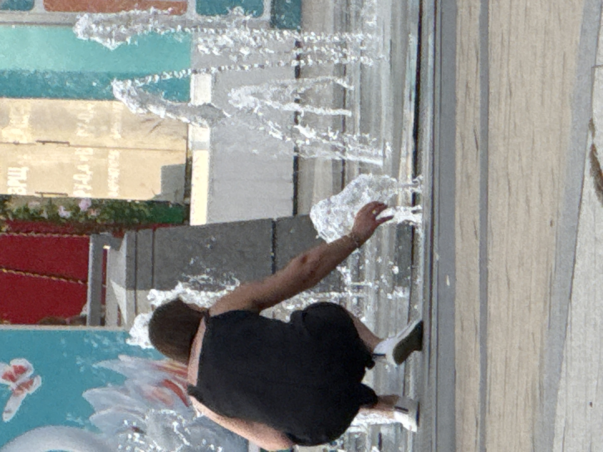 In Zelenograd, Russia, a person is seen interacting with a fountain. The subject, wearing a black shirt and shorts with white sneakers, is reaching out towards the water jets, possibly to feel the spray or catch some water. The scene appears to be outdoors, with a tiled or concrete ground surface and what seems to be a decorative wall or structure in the background. The wall has sections of different colors, including turquoise and beige, with some text visible on the beige sections which is difficult to decipher fully due to image resolution. One cropped section shows what might be Russian text like "ЕРЦ".
The water from the fountain is captured mid-air, creating a dynamic and refreshing element in the image. The lighting suggests it's daytime, likely a sunny day given the brightness and the water's reflection. There are no strong indicators of specific weather conditions beyond the presence of the fountain, which implies a temperate or warm climate. The overall mood seems relaxed and playful, with the subject enjoying the cooling spray of the fountain on a warm day. The presence of a blue painted area with white floral motifs in the lower part of the image suggests a public recreational area or a park.