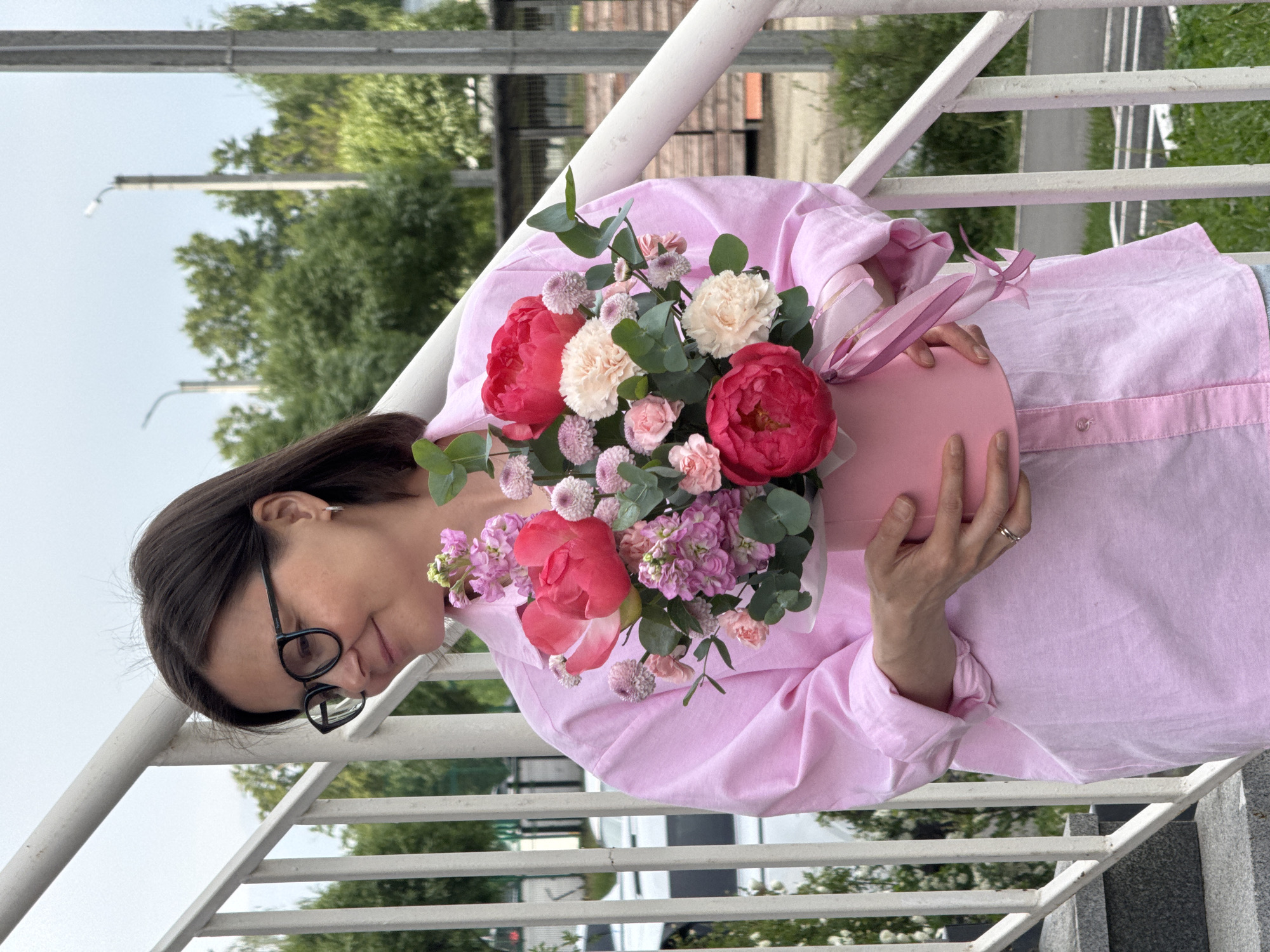 A woman with dark hair and glasses stands on a metal railing outdoors, holding a pink box filled with flowers. The flower arrangement consists of pink peonies, white carnations, eucalyptus leaves, and other small pink blooms. The box has a pink ribbon tied around it. The woman wears a light pink collared shirt.
The background includes trees and a building. The location is Izhevsk, Russia.