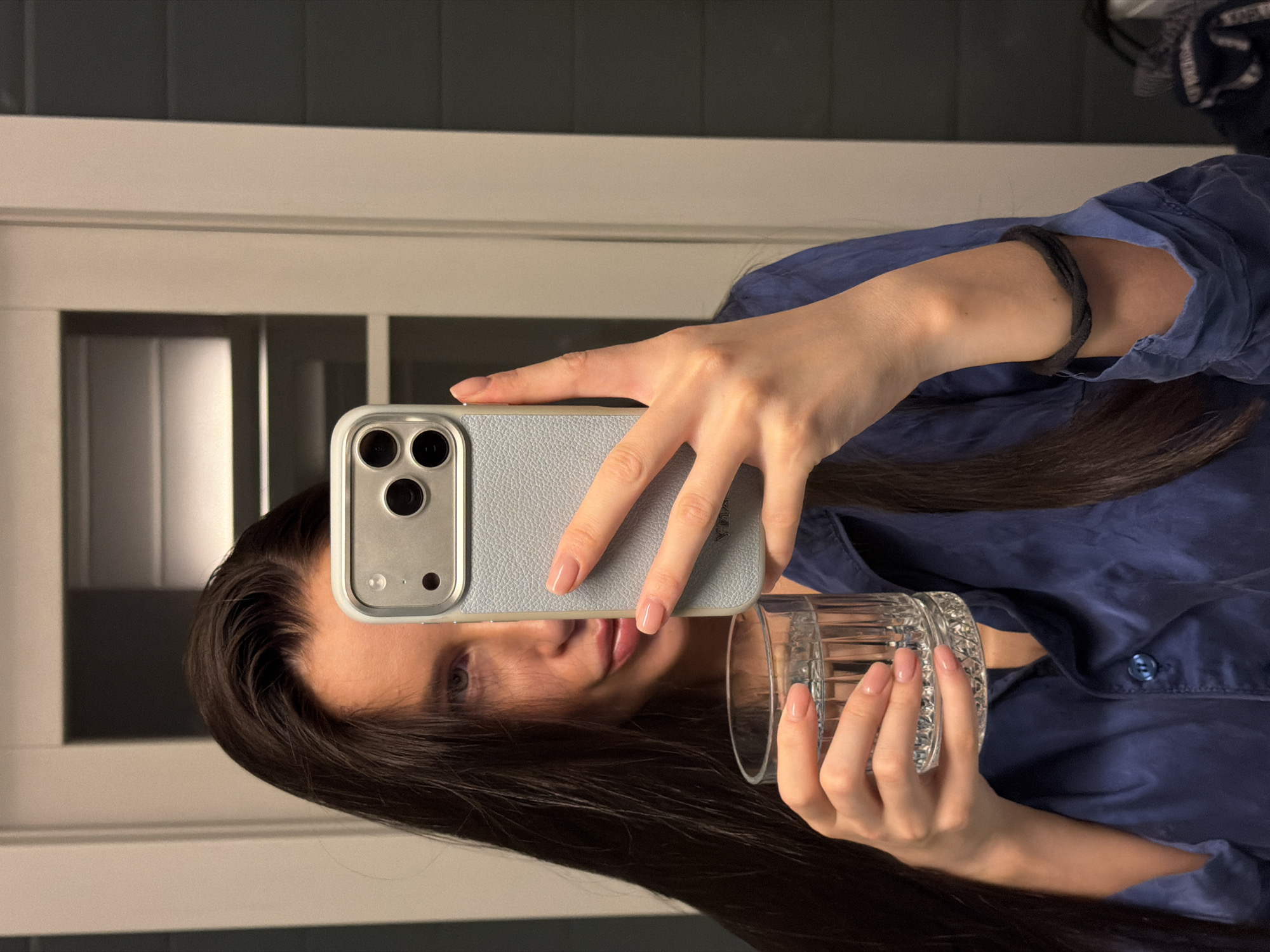 An indoor, vertically oriented image captures a person taking a mirror selfie (or a selfie using a reflective surface) in what appears to be a domestic setting in Khimki, Russia.
The primary subject is a person, likely female, with long, dark hair. Their face is partially visible in the reflection on the phone or a mirror behind it, showing one eye and parts of the nose and mouth. They are wearing a blue button-up shirt.
In their right hand, the person holds an iPhone with a light blue, textured case and a silver-colored frame around the triple-lens camera system. Their fingers, adorned with light pink/nude nail polish, are wrapped around the phone. A black elastic hair tie is worn on their right wrist. Their left hand holds a clear, cut-glass style tumbler. The fingernails on this hand also feature light pink/nude polish.
The background consists of an interior space with light-colored horizontal architectural trim and darker vertical elements, suggesting walls or furniture. The lighting is soft and even, indicating an indoor environment. There is no visible text in the image. The overall scene suggests a casual, personal moment.