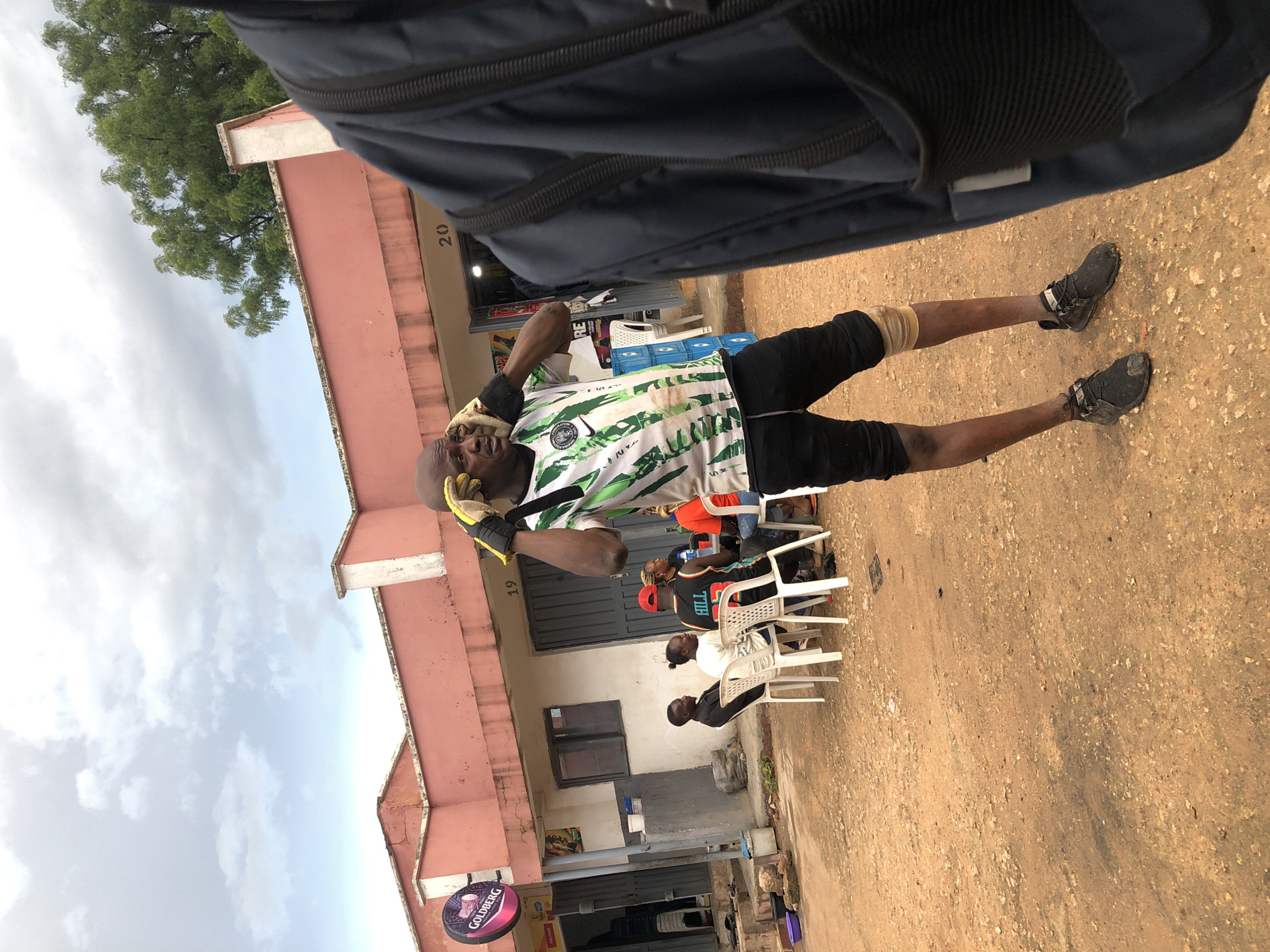 The image depicts an outdoor scene in Jos, Nigeria, with a focus on a man in the foreground wearing a green and white Nigerian soccer jersey and black shorts. He is holding his hands to his head, seemingly adjusting something over his ears, and appears to be wearing gloves. A knee brace is visible on his right leg. The background reveals a building with pink walls and white trim, featuring several storefronts and what appear to be seating areas outside. Several people are seated at white chairs, some of whom are engaged in conversation. A Goldberg sign is visible on one of the storefronts. The ground is covered in dirt and small stones, indicative of an unpaved area. The sky is overcast. The perspective suggests the photo was taken by someone standing close to the man in the foreground, with part of a dark backpack visible on the right side of the frame.