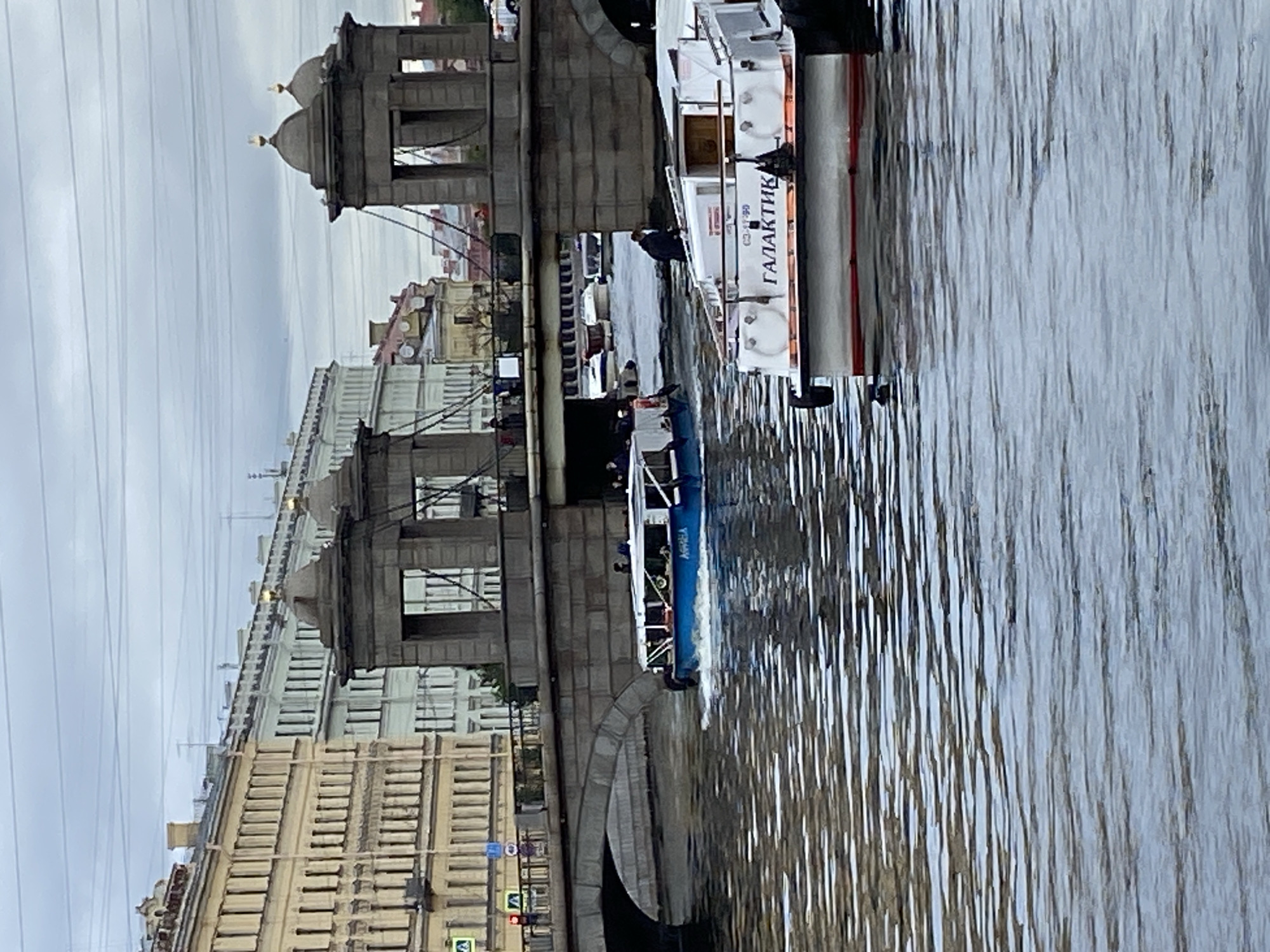 A scene of a canal in Saint Petersburg, Russia, with boats and buildings is depicted. In the foreground, a white boat with a red stripe along its waterline is docked. On its side, written in Cyrillic, is the name "ГАЛАКТИК" (Galaktik). A person in dark clothing is standing on this boat. Behind it, a blue and white boat with passengers is moving through the canal. Further in the background, a stone bridge with two domed towers spans the canal. On the far bank of the canal, there are multi-story buildings with classical architecture, characteristic of Saint Petersburg. The sky is overcast, and the water is choppy, reflecting the muted light.
