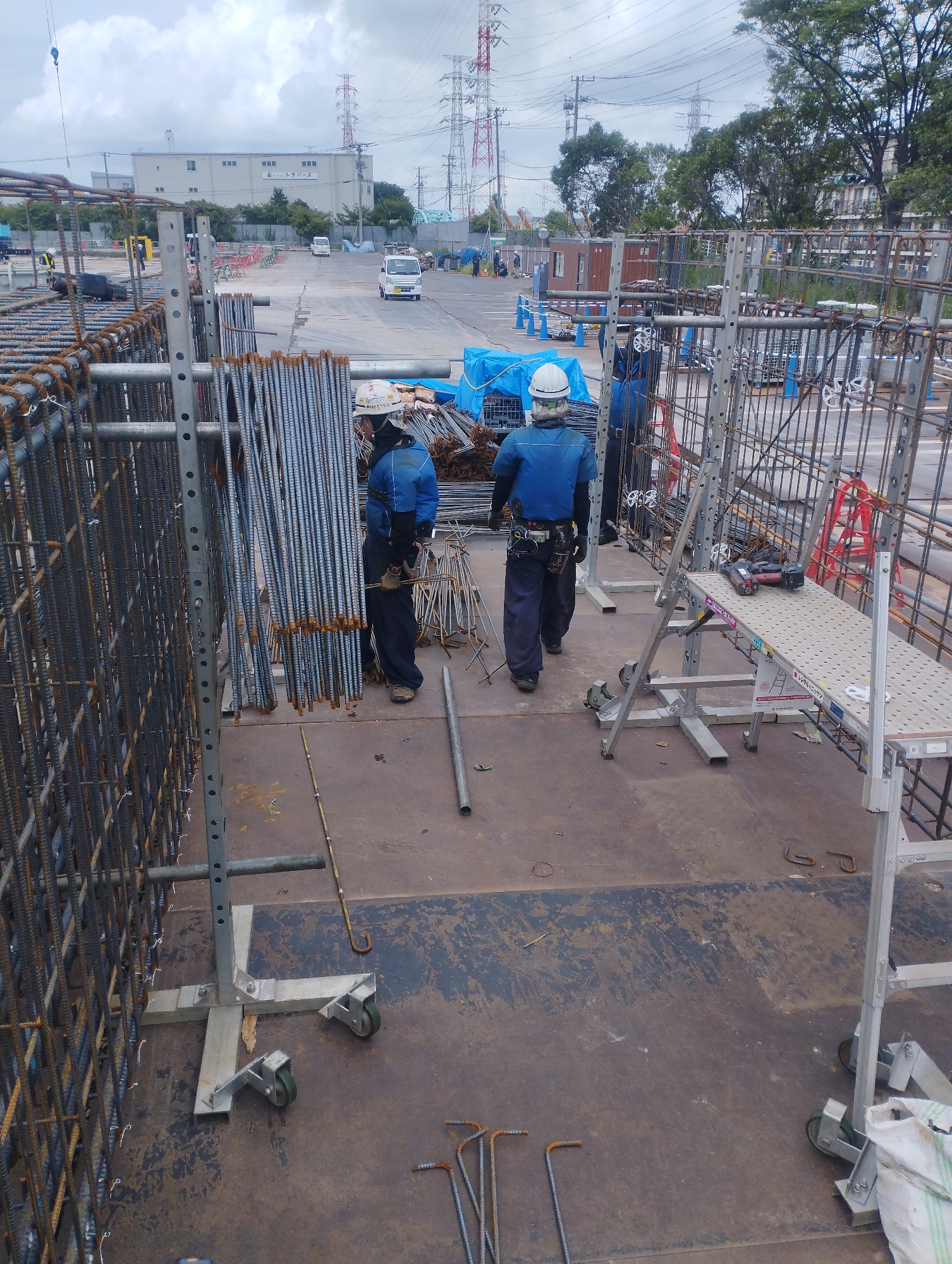 The image captures an outdoor construction site in Ichikawa, Japan, likely a part of the Tokyo metropolitan area, as indicated by the infrastructure and style of buildings visible in the background. The foreground is dominated by rebar structures and scaffolding, suggesting ongoing civil engineering or building work.

Two construction workers, dressed in blue uniforms and white hard hats, are visible. The worker on the left is facing away from the camera, with their back to the viewer, and appears to be standing or walking on the scaffolding. The worker on the right is also facing away, with their body angled slightly, looking towards a pile of rebar. Both workers are equipped with safety harnesses and tools, including gloves and what appear to be binding machines on the scaffolding.

The environment is characterized by a cloudy sky, with power lines and industrial buildings in the distance. Several white vans are parked or driving on a paved road in the middle ground. The ground surface is a mixture of concrete and darker, possibly asphalt or worn-down material, with scattered debris. Other construction materials, such as tarpaulin-covered items and more rebar, are present. A rolling scaffold platform with a power tool is positioned in the mid-right of the frame. The overall impression is a busy, functional construction zone.