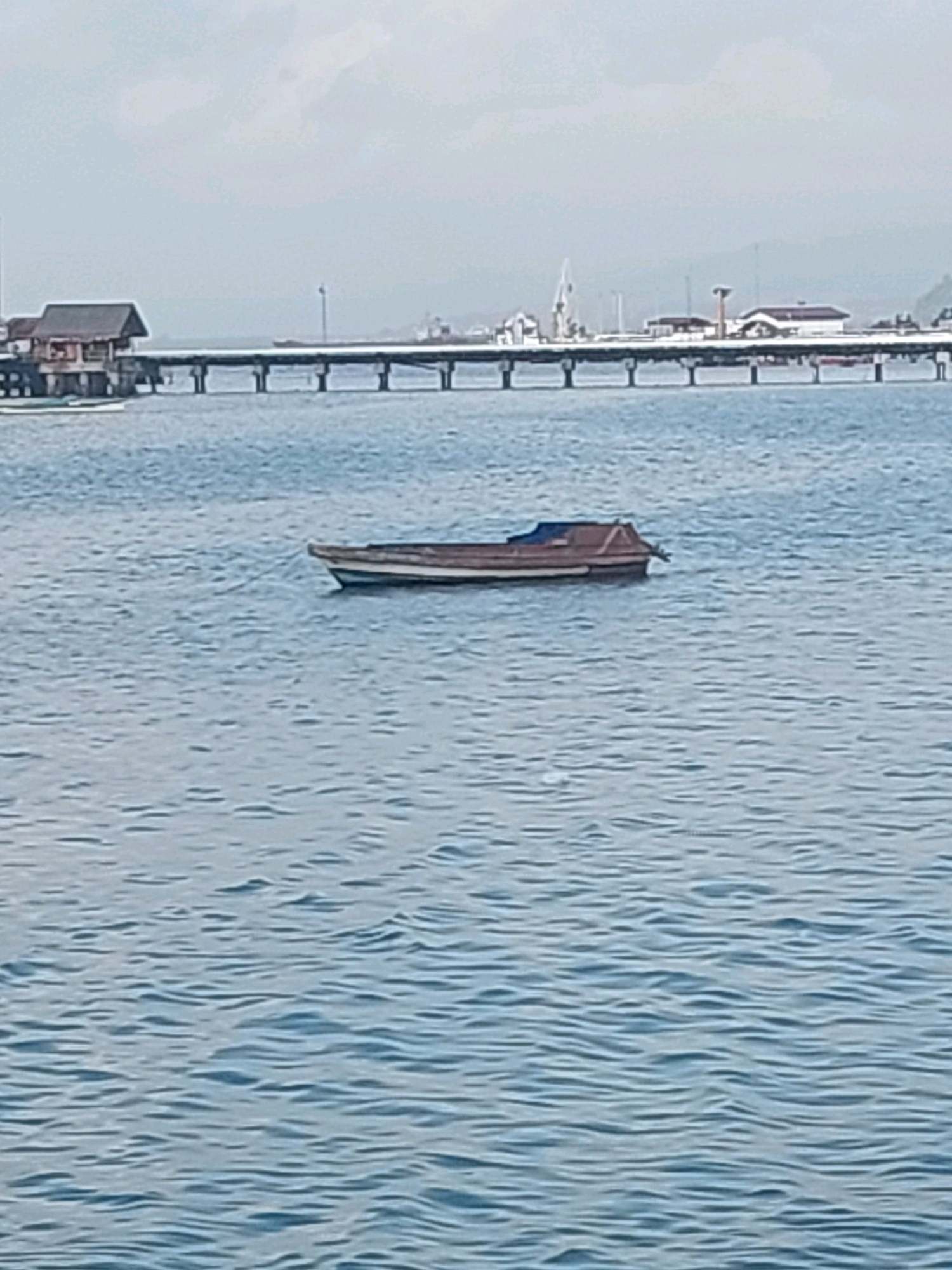 The image shows a calm sea with a small wooden boat floating in the foreground. In the background, a long pier stretches across the water, connecting to a building on stilts. The sky is overcast, suggesting a possibly humid or rainy day. The water appears to be a deep blue, with small ripples on the surface. The pier is supported by numerous pilings and has railings along its length. The building on the pier has a dark roof and several windows. Further in the background, the outline of a city or town can be seen, with structures and possibly cranes or industrial equipment visible. The overall scene suggests a coastal area, possibly a harbor or a fishing village. The city of Ambon, Indonesia, is known for its beautiful harbors and coastal landscapes, making this scene consistent with that location.