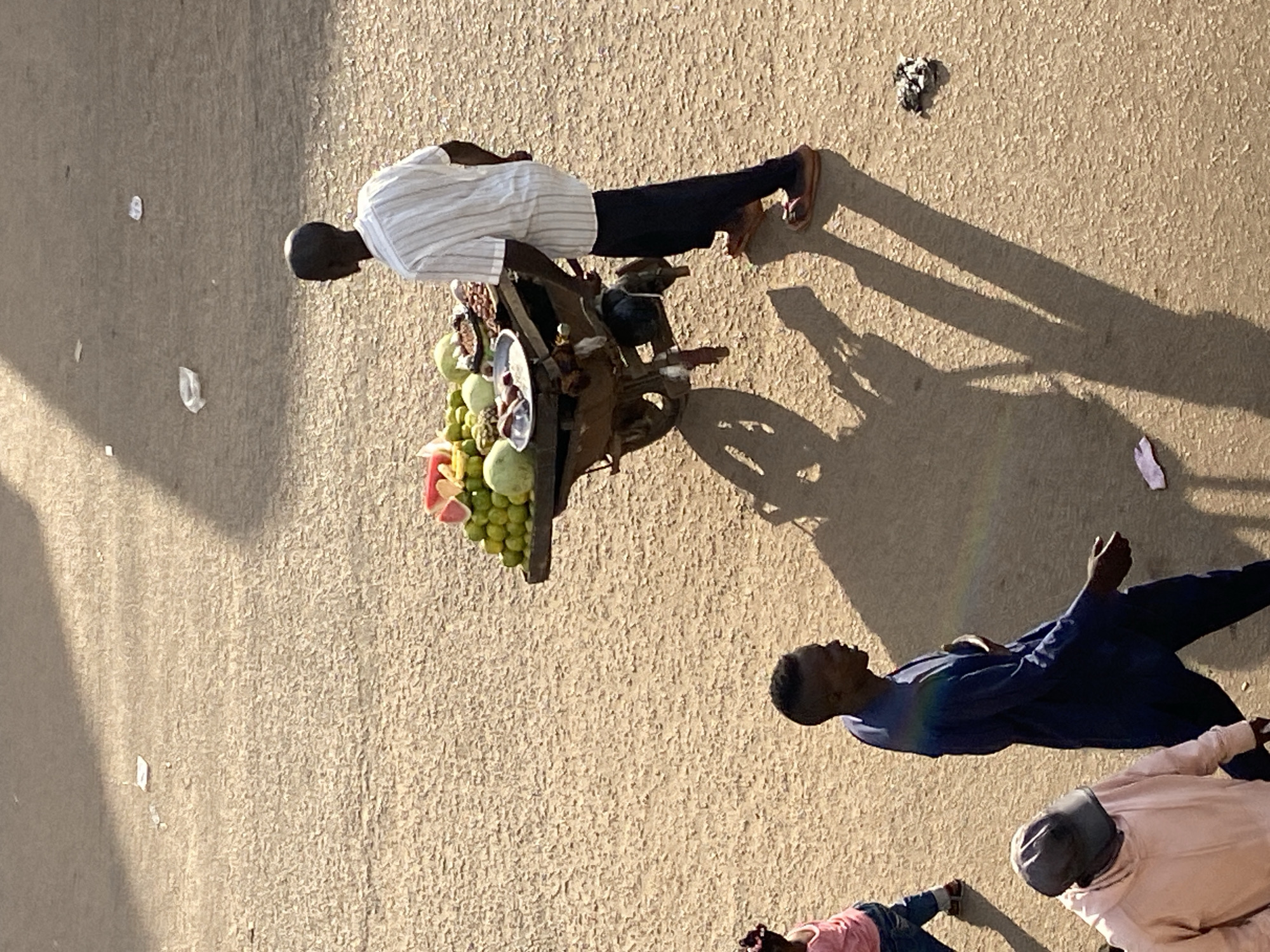 This image captures an overhead view of a street scene in Bukuru, Nigeria, under strong natural lighting conditions. The ground surface is a textured light brown, consistent with unpaved earth or coarse asphalt.
The primary subject is a dark-skinned male pushing a two-wheeled wooden cart laden with produce. He is wearing a vertically striped white and grey short-sleeved shirt, dark trousers, and dark sandals. He faces away from the camera, moving towards the upper-right of the frame. The cart holds a variety of fruits, including a stack of green spherical items (possibly limes or small melons), a cut watermelon with exposed red flesh, and a metallic bowl containing darker items, possibly nuts or dried fruits. The man's shadow, along with the cart's shadow, extends long and dark towards the lower-left of the frame, indicating the sun's position.
Three other individuals are visible. In the mid-lower-left, a dark-skinned male walks towards the lower-left, wearing a dark blue long-sleeved tunic (agbada style). His head is turned slightly to his right. A faint rainbow-like chromatic aberration is present on his back. To his left, a young child, partially visible, wears a pink top and blue jeans, walking in the same general direction. In the lower-left corner, another individual is partially visible, wearing a light pink hooded sweatshirt and a dark baseball cap that obscures their face.
Scattered across the ground are several pieces of white litter, appearing to be paper or plastic, and a small clump of dark debris in the lower-right quadrant. The overall scene depicts a busy street or market environment.
