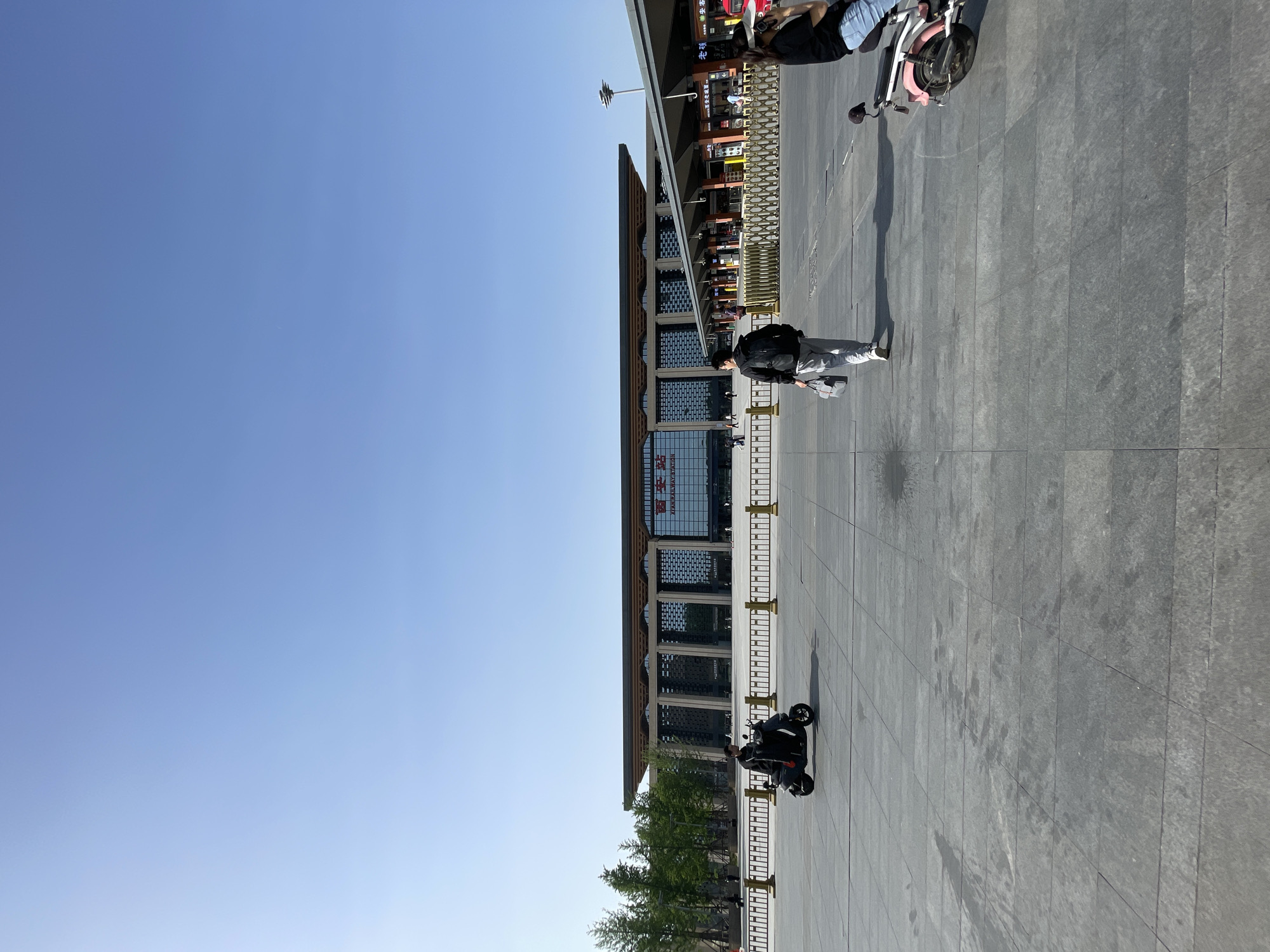 A ground-level, vertically oriented photograph shows a wide paved plaza in Xincheng District, China, under a clear, bright blue sky. A modern building with a glass facade and horizontal architectural elements runs horizontally across the middle of the image, with its base near the bottom edge.
A person wearing grey clothing and carrying a backpack walks across the plaza, away from the building. Closer to the building, a black motorcycle sits parked. Further away, closer to the top edge of the image, are two other people, one with a pink electric bike. A thin row of trees and shrubs can be seen along the bottom edge of the photograph, in front of the building. The paving appears to be made of large, square tiles. A barricade runs parallel to the front of the building.