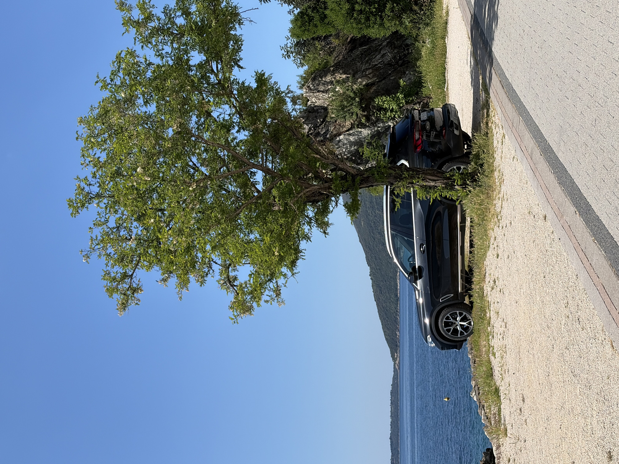 The image depicts a black SUV parked on the side of a road, partially shaded by a tree with green foliage and some white blossoms. The road is made of gravel and bordered by a gray concrete curb. To the right of the car, there is a sharp drop to the sea, with a yellow buoy visible in the water. Behind the car, there's a rocky cliff covered in vegetation. In the background, a mountain ridge can be seen across the water. The sky is clear and blue. While the exact location within Herceg Novi cannot be determined solely from the image, the geography and vegetation suggest a coastal road in that region of Montenegro.