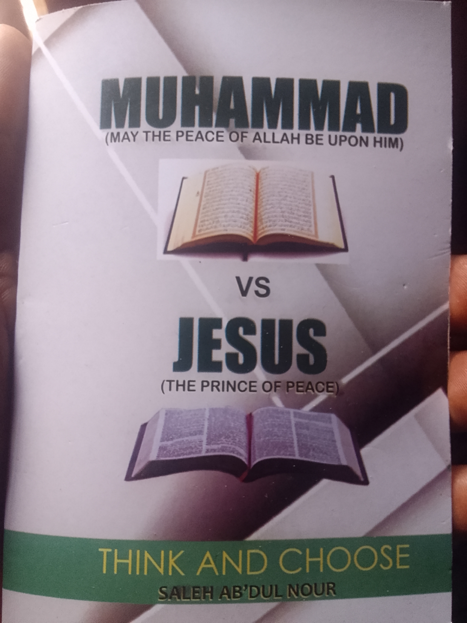 A person's hand holds a book with the title "Muhammad vs. Jesus." The book has pictures of open books, presumably religious texts, positioned above the names of the religious figures. Underneath, it says "Think and Choose" followed by the author's name, "Saleh Ab'dul Nour."