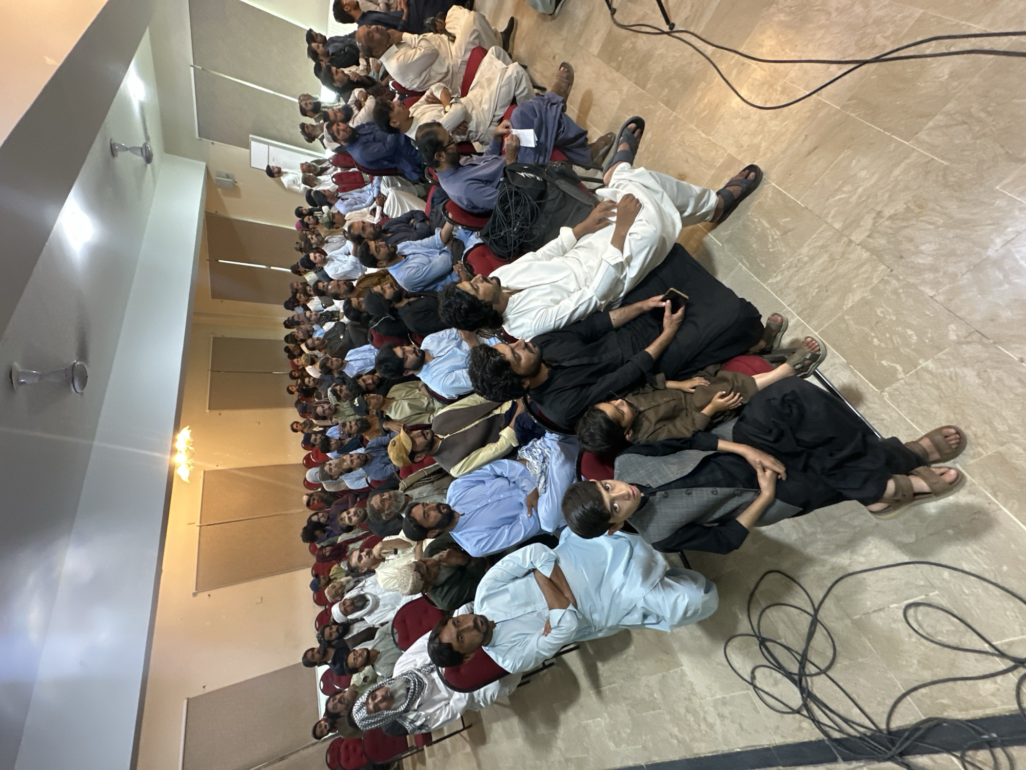 The image shows a large group of men seated in rows in what appears to be an indoor hall or auditorium. The men are dressed in traditional Pakistani clothing, with many wearing white or light-colored shalwar kameez and some wearing head coverings. The rows of chairs are densely packed, and many of the men are looking towards the front of the room, suggesting they are attending a lecture or presentation. The image was taken in Quetta City Tehsil, Pakistan.
