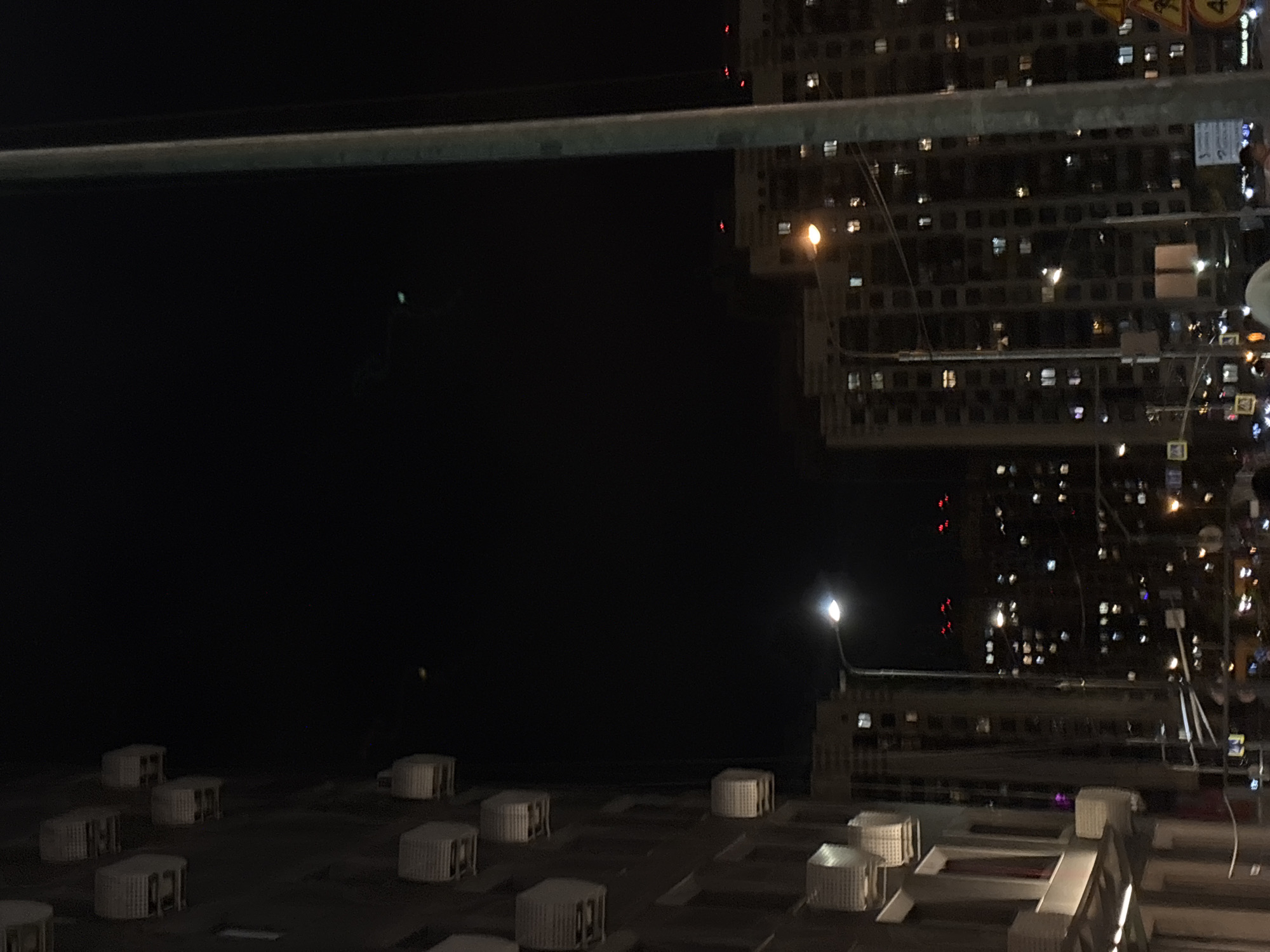 Captured from a rooftop in Vnukovo, Russia, at night, the image shows an urban scene. The rooftop itself is occupied by several rectangular structures, possibly air conditioning units. The background is dominated by a large, illuminated apartment building with a grid of windows. A street lamp is seen near the building. The sky is dark and unclear. A horizontal bar is visible in the top-left corner.