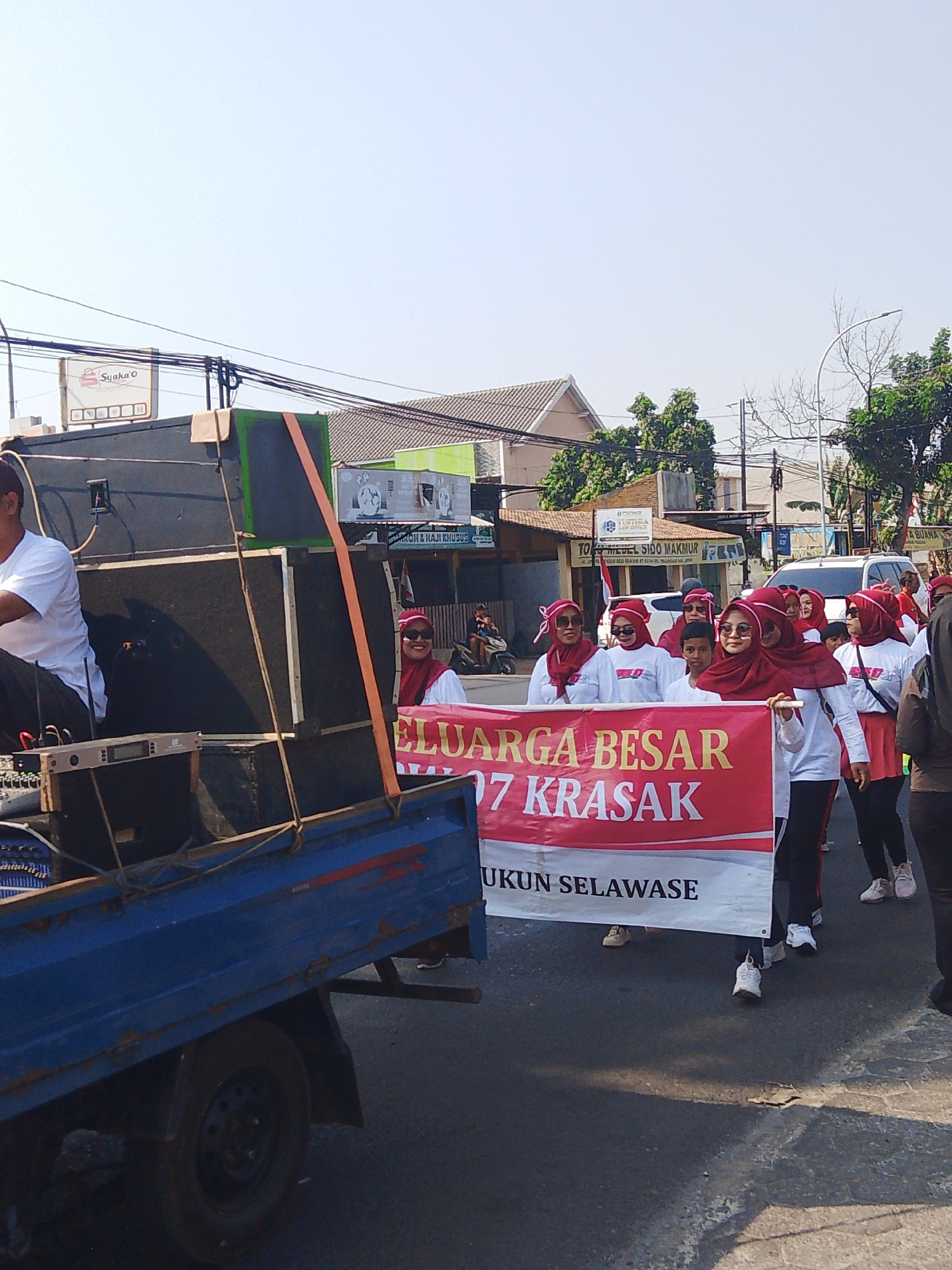 A group of people, primarily women wearing red headscarves and white shirts, are participating in a parade on a street in Rejosari, Indonesia. They are holding a large red and white banner that reads "KELUARGA BESAR RT 07 KRASAK" and "UKUN SELAWASE". Behind them, a flatbed truck is visible, loaded with large speakers. The truck is blue and shows signs of wear. The background features buildings, trees, and utility poles, indicating an urban or semi-urban setting. The sky is clear and bright, suggesting daytime. The overall scene depicts a community event or procession.