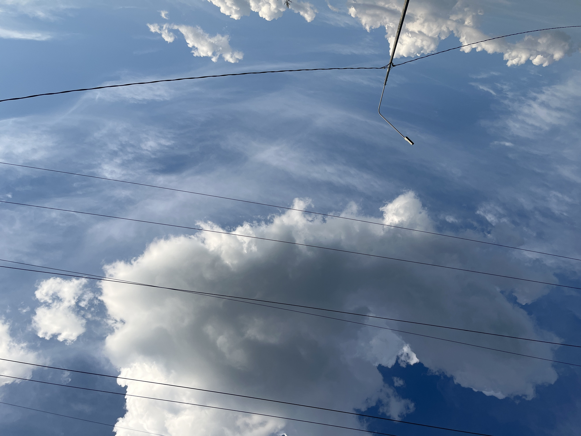 The image depicts a bright, clear sky with scattered clouds, likely taken in Novogusel'skii, Russia. Several dark power lines stretch horizontally across the frame, some appearing taut and others with a slight sag. In the lower left quadrant, a silver lamppost with a sleek, modern design rises vertically, its curved arm extending towards the right. In the distance, smaller, fluffy white clouds are visible near the horizon, contrasting with a large, dense, gray-white cloud formation dominating the upper portion of the sky. The sky transitions from a deep blue at the top to a lighter, more translucent blue towards the bottom.