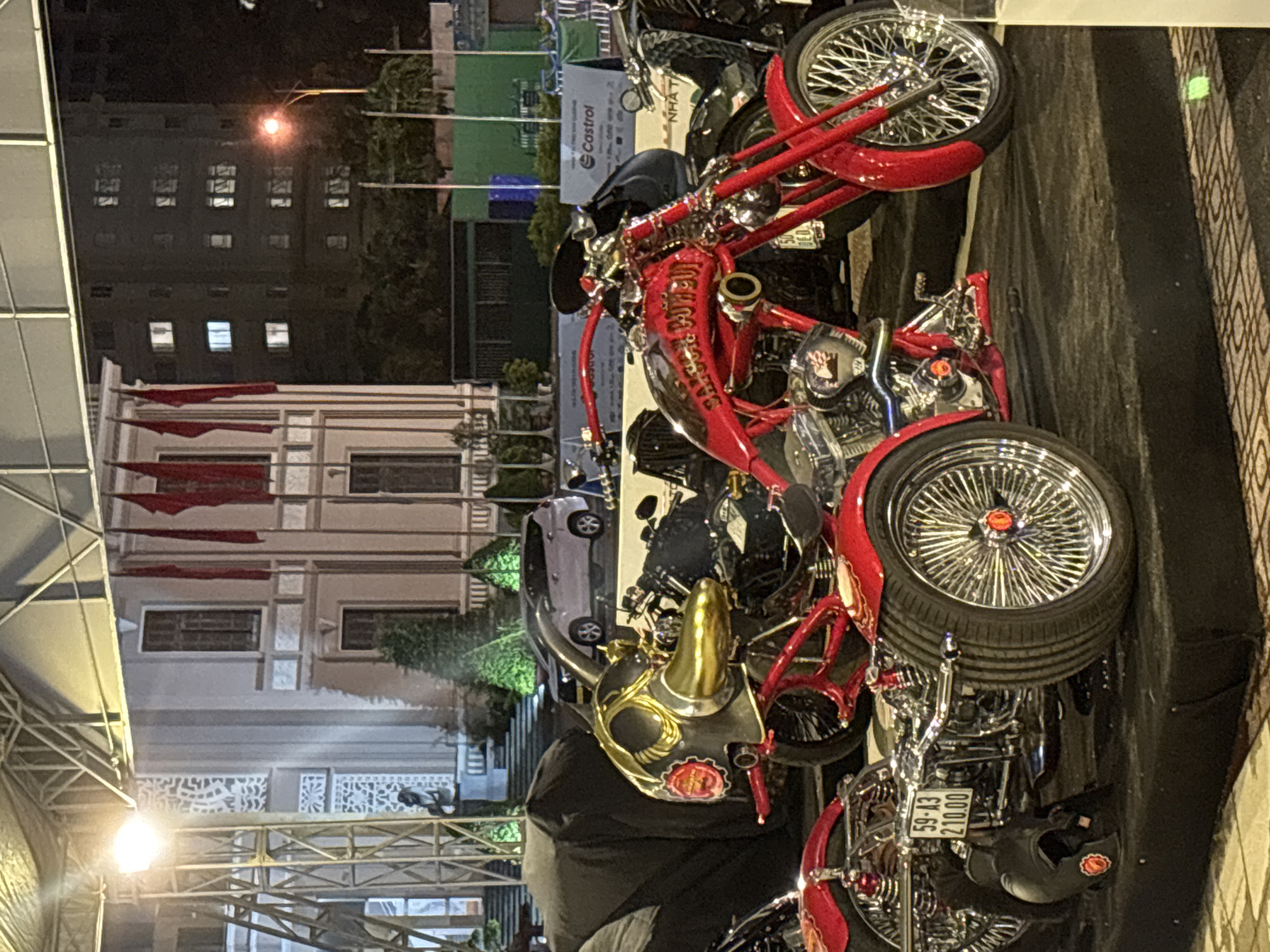 Captured indoors, the photograph showcases a motorcycle. The bike boasts a vibrant red frame and a chrome engine, wheels, and handlebars. Its front wheel is notably large, with a high, curved red fender. The rear wheel, smaller, also features a chrome fender.
The background presents a multi-story building with numerous windows. Red flags adorn the building, suggesting a national or local event. The setting appears to be Nha Trang Ward, Vietnam. The overall lighting is dim, with light fixtures contributing to the indoor illumination. The image is rotated counter-clockwise.