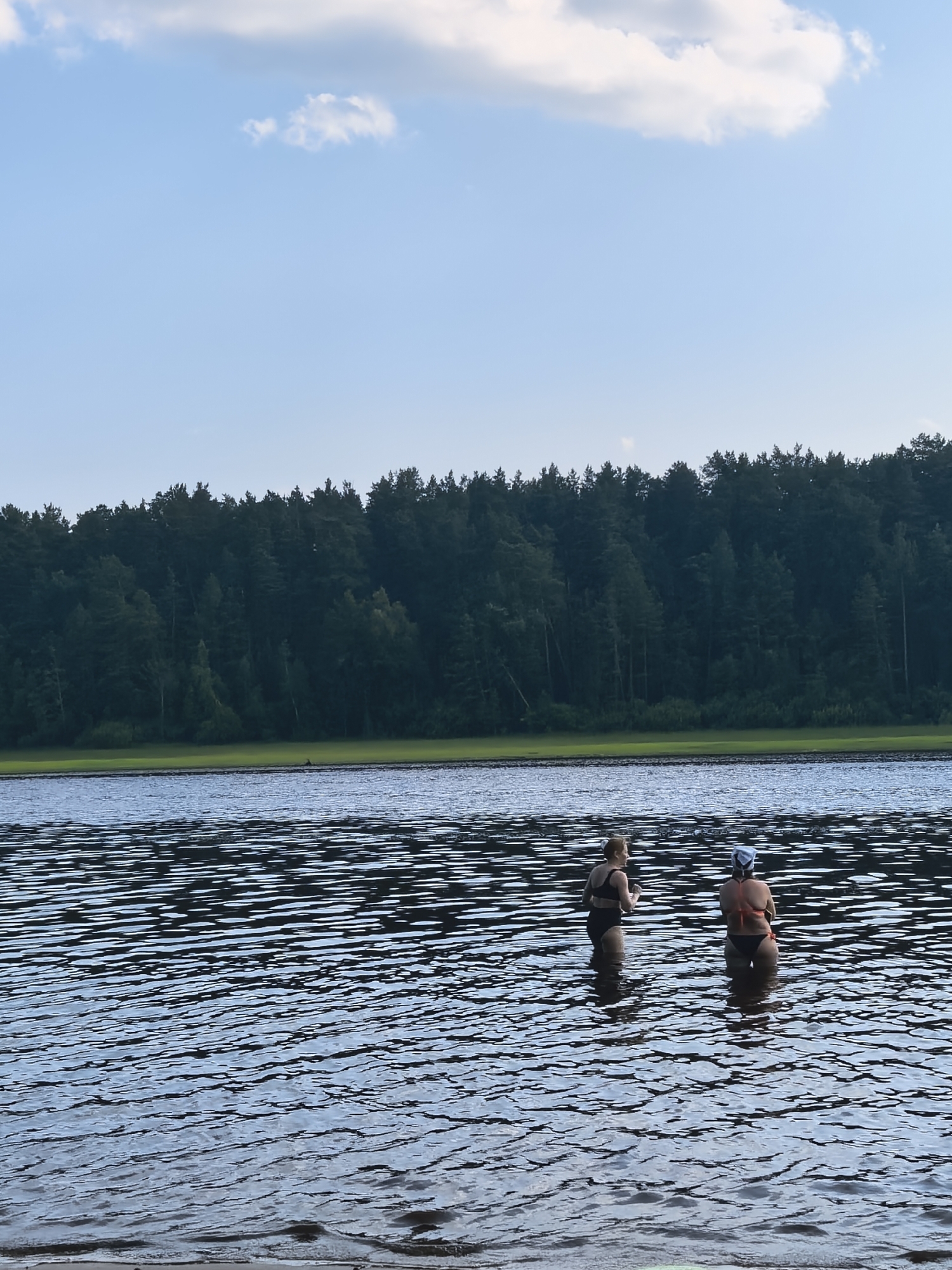 Two people stand in the shallow waters of a lake, facing away from the camera. The person on the left wears a black bikini and has blonde hair pulled back. The person on the right wears a bikini with an orange top and black bottoms and has a light-colored swim cap on. The water is rippled and reflects the sky. In the background, a dense, dark green forest lines the opposite shore of the lake. Above the forest, a light blue sky is visible with scattered white clouds. The scene appears to be set during daytime, likely late afternoon or early evening, given the soft light. The location is identified as Sysert, Russia. There is no visible text in the image.