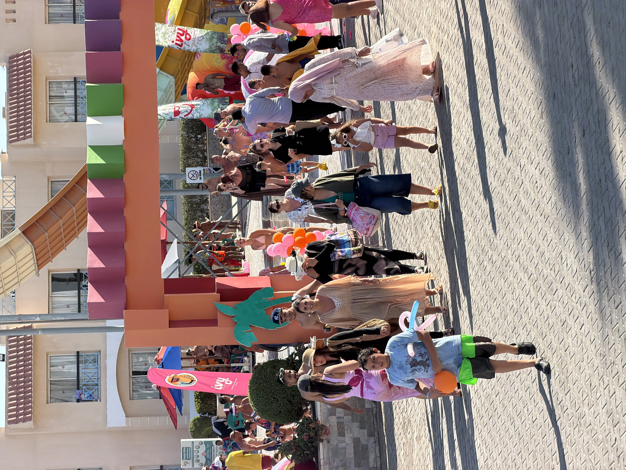 The image captures a sun-drenched outdoor scene at a water park or resort in Sharm El Sheikh, Egypt, on a paved walkway. A group of people, varying in age and attire, are walking and interacting. In the foreground, a young boy in a light blue t-shirt and dark shorts holds an orange balloon and a blue balloon animal. To his right, a woman in a light-colored, flowing dress with a floral pattern walks with a shopping bag. Behind them, several individuals are seen, including a woman in a straw hat and a black cardigan, and another in a beige dress.
The background reveals parts of the resort's architecture, including multi-story buildings with balconies and windows, and colorful water slides and structures indicative of a recreational facility. Brightly colored banners and umbrellas are visible, adding to the vibrant atmosphere. The lighting suggests it is daytime, with strong shadows cast on the patterned pavement. The overall impression is of a lively, tourist-oriented environment.