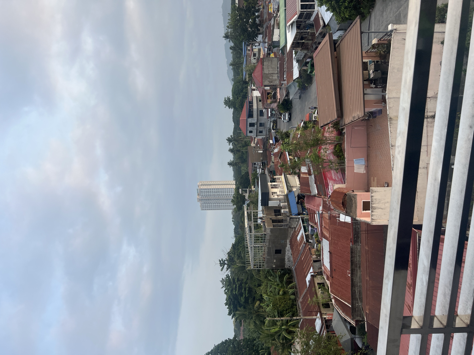 This is an aerial view of a residential area in Davao City, Philippines, under a cloudy sky. The scene features a dense collection of houses with predominantly corrugated iron roofs, many of which are rusted, indicating an older, possibly less affluent, neighborhood. A multi-story building under construction with a metal frame is visible in the mid-ground, suggesting ongoing development. In the background, a tall, modern high-rise apartment building stands out against the lush green trees and distant hills, creating a contrast between older and newer urban development. A paved road winds through the community, with a few parked vehicles and a couple of individuals walking, indicating normal daily activity. The foreground is partially obstructed by a metal railing, suggesting the photograph was taken from a balcony or elevated vantage point.