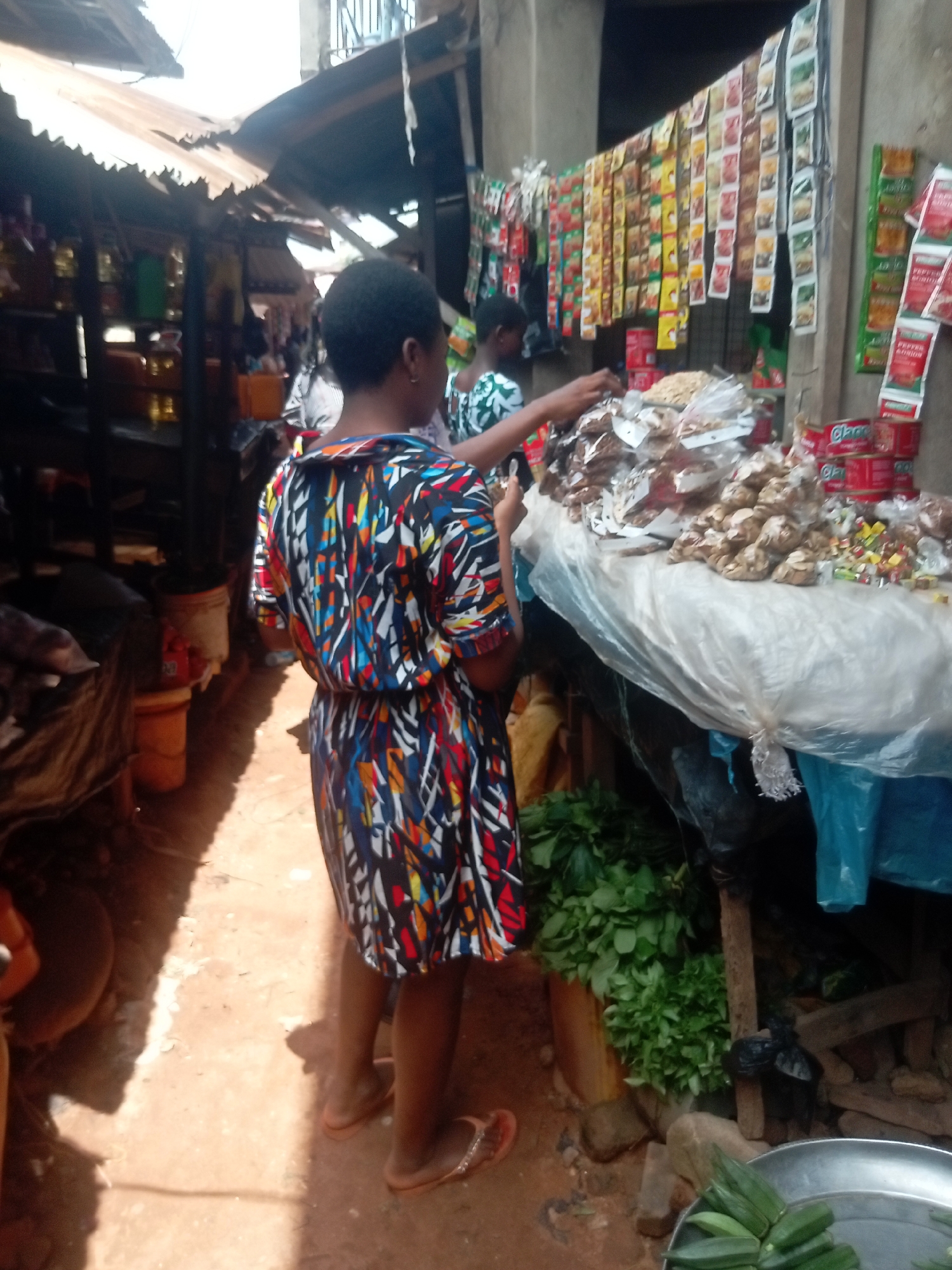 The image shows a bustling market scene in Ogidi, Nigeria. A woman wearing a colorful patterned dress and flip-flops stands on a dirt floor, facing a vendor's stall. The stall is covered with a white plastic sheet and displays an assortment of goods, including packaged nuts, candies, and canned goods. A variety of spices are hung on the wall behind the stall. The vendor, partially visible, is reaching for an item on the stall. Beneath the stall, there is a pile of fresh green leafy vegetables and a silver tray containing okra. The market is covered by a makeshift roof constructed from corrugated metal sheets. Other people are visible in the background, moving through the crowded market area.