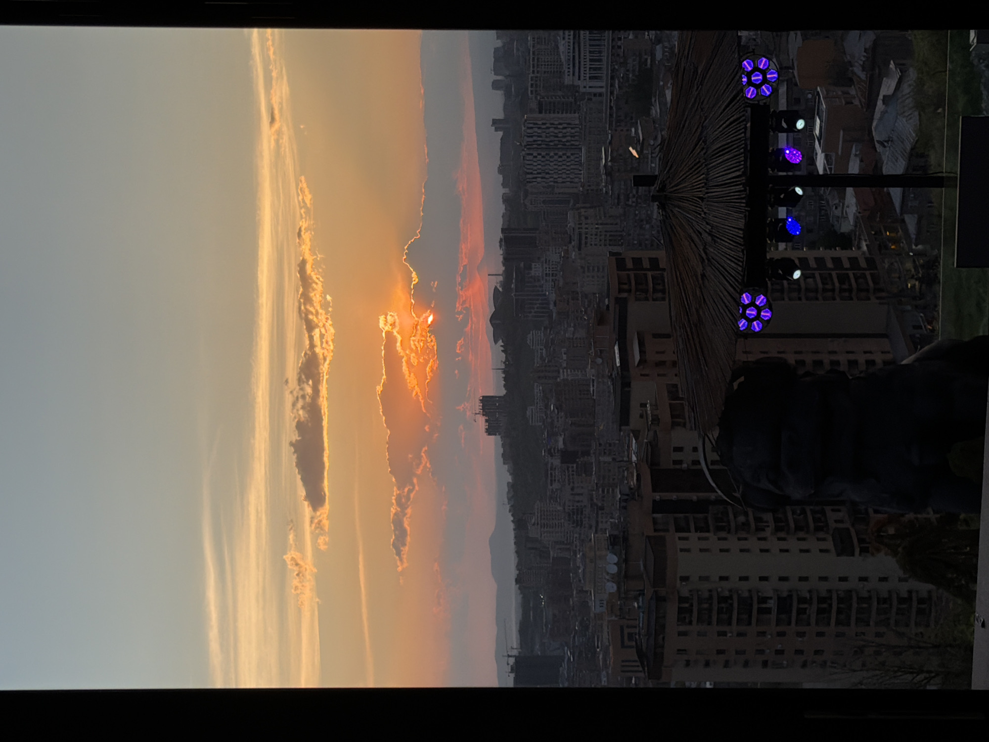 The image captures a vibrant sunset or sunrise over the city of Yerevan, Armenia, viewed from an elevated perspective, likely a rooftop. The sky dominates the upper portion of the frame, showcasing a dramatic transition of colors from a soft blue at the top to brilliant oranges, yellows, and reds near the horizon. The sun is partially obscured by a thick band of clouds in the mid-left, casting intense golden rays and highlighting the intricate textures of the clouds. Streaks of light and warmer hues extend across the sky.
Below the colorful sky lies a dense cityscape, rendered mostly in dark silhouettes against the brighter light. Numerous buildings, both residential and commercial, fill the urban landscape, stretching towards faint, hazy mountains in the distance. Several construction cranes are visible, indicating urban development.
In the right foreground, a large, dark structure resembling a thatched umbrella or canopy is prominent. Attached to a vertical pole near this canopy are multiple stage lights or decorative lights. Some of these lights are illuminated, emitting distinct circular patterns of purple and blue light. A dark, indistinct shape, possibly a person or part of a seating area, can be seen in the bottom right corner, suggesting a social gathering spot.
The overall atmosphere is one of serene beauty, capturing the dramatic light of the golden hour over a bustling city as evening begins or morning breaks. There is no visible text in the image.