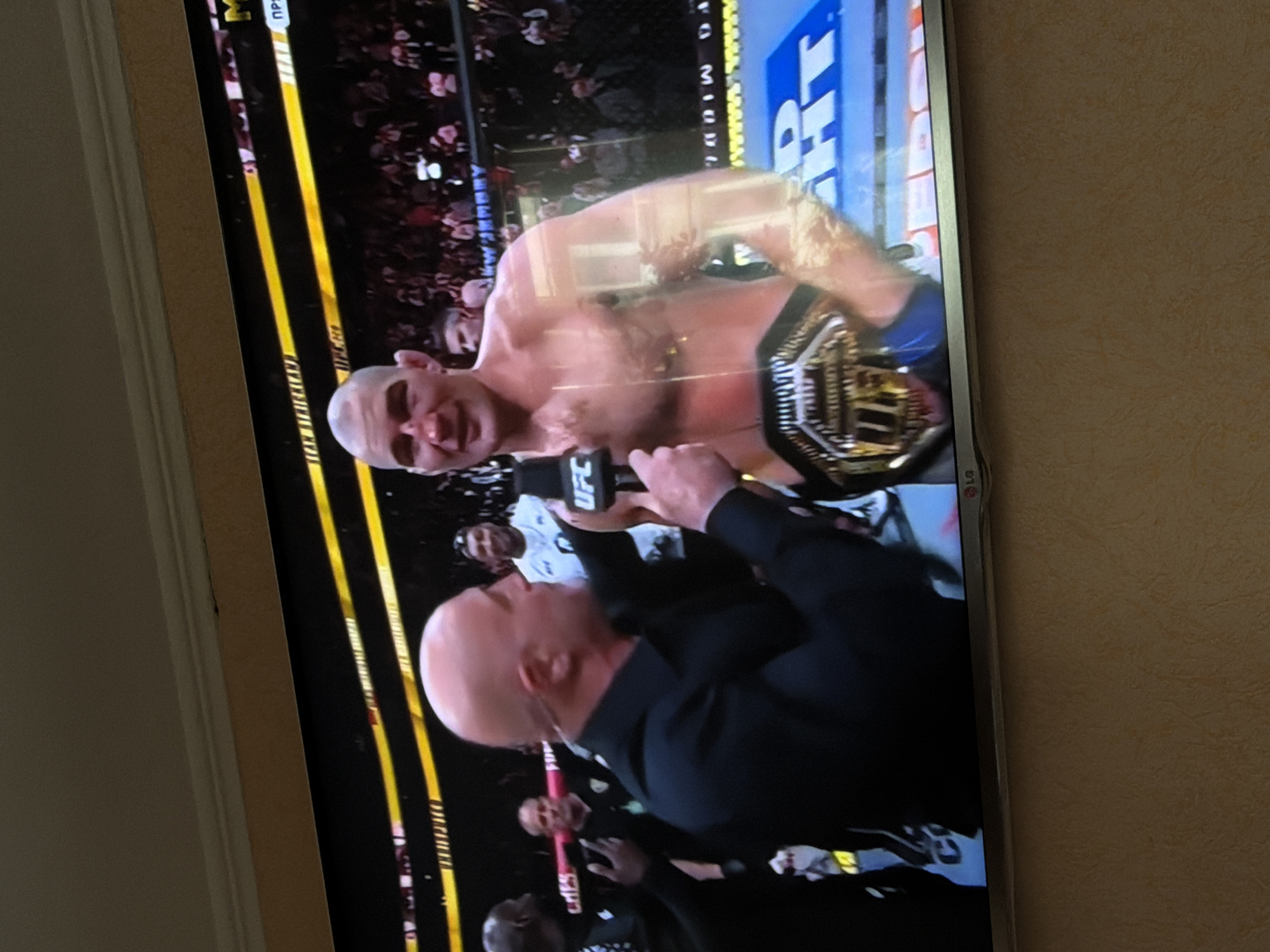 A photograph shows a television screen displaying a scene of a UFC fighter with a championship belt, being interviewed. The interviewer has a bald head. The fighter appears to be in the center of the ring with spectators in the background. The photograph was likely taken indoors, evidenced by the beige wall visible to the right and the TV frame to the left. There is no verifiable connection to the city of Beryozovka, Russia, based on the available visual information.