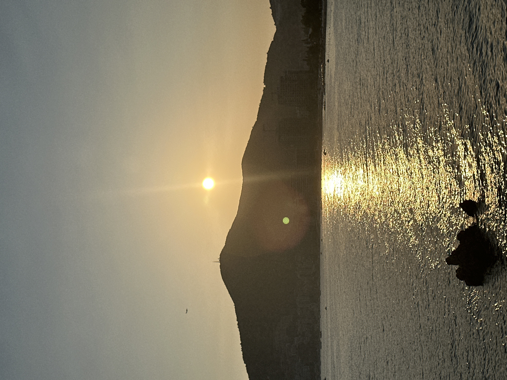 The image depicts a serene sunset over the sea with the silhouette of a mountain range in the background, likely Budva, Montenegro. The sun, a bright orb, is positioned in the upper center of the frame, casting a golden, shimmering reflection on the water's surface. The mountain range, dark and rugged, dominates the horizon, with subtle signs of buildings and infrastructure built into its slopes, suggesting a coastal city. A few small boats are visible on the water, scattered across the expanse. The sky transitions from a pale blue at the top to a warm, golden hue near the horizon, indicative of the twilight hour. In the upper left corner of the sky, a small, dark silhouette of an airplane is visible. The foreground features a dark, rocky outcrop emerging from the water, adding a natural element to the scene.