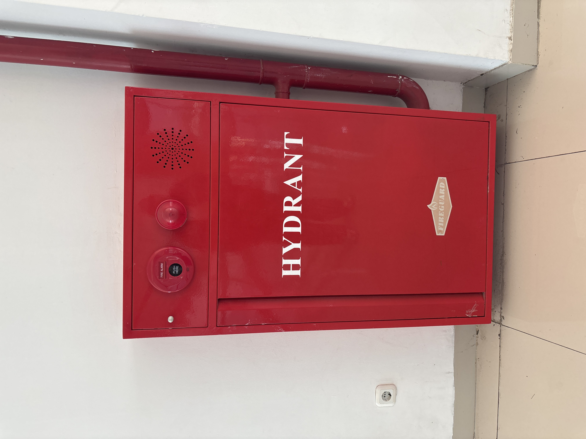 A red fire hydrant cabinet is mounted on a white wall in Cirebon, Indonesia. The cabinet has a door with the word "HYDRANT" in white lettering and a "FIREGUARD" logo at the bottom. Above the door, there is a fire alarm button and a speaker grille. A red pipe runs vertically alongside the cabinet. A white electrical outlet is visible on the wall to the left of the cabinet. The floor is tiled in a cream color.