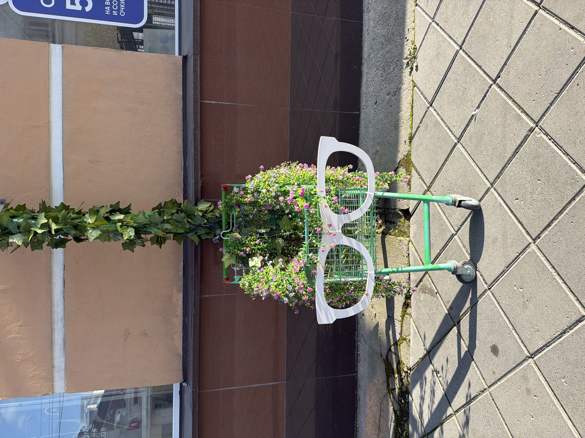 A storefront displays a shopping cart decorated with artificial ivy and small pink and purple flowers, resembling a vertical garden. Attached to the front of the cart is a large, white, cut-out of eyeglasses. The cart is positioned on a paved sidewalk in front of a building with a light brown and dark red facade. Shadows from the cart and surrounding structures are cast across the pavement. Text on the building's facade, visible through a window on the left, appears to be in Russian and includes the word "МАГАЗИН" (shop). A blue sign with white text on the right side of the building also appears to be in Russian. The overall scene suggests an outdoor display outside a commercial establishment in Yekaterinburg, Russia.