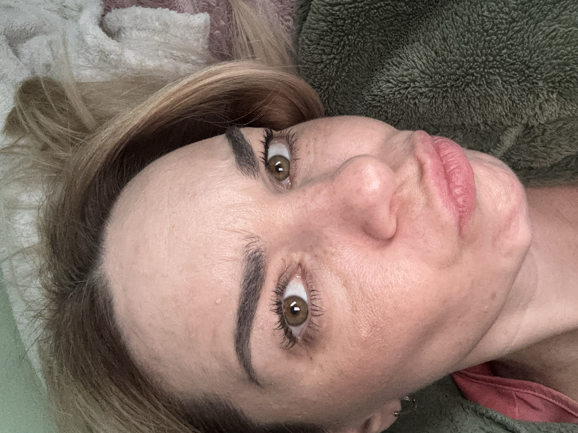 A close-up shot of a Caucasian woman lying down. Her head is tilted at an angle, presenting a direct view of her face. She has light brown hair, prominent eyebrows, and hazel eyes. Her skin is fair, and there are visible lines around her eyes and on her forehead. Her lips are slightly pursed. She is wearing a red top with an unidentified green and pink blanket in the background. The location is Unknown, Russia.
