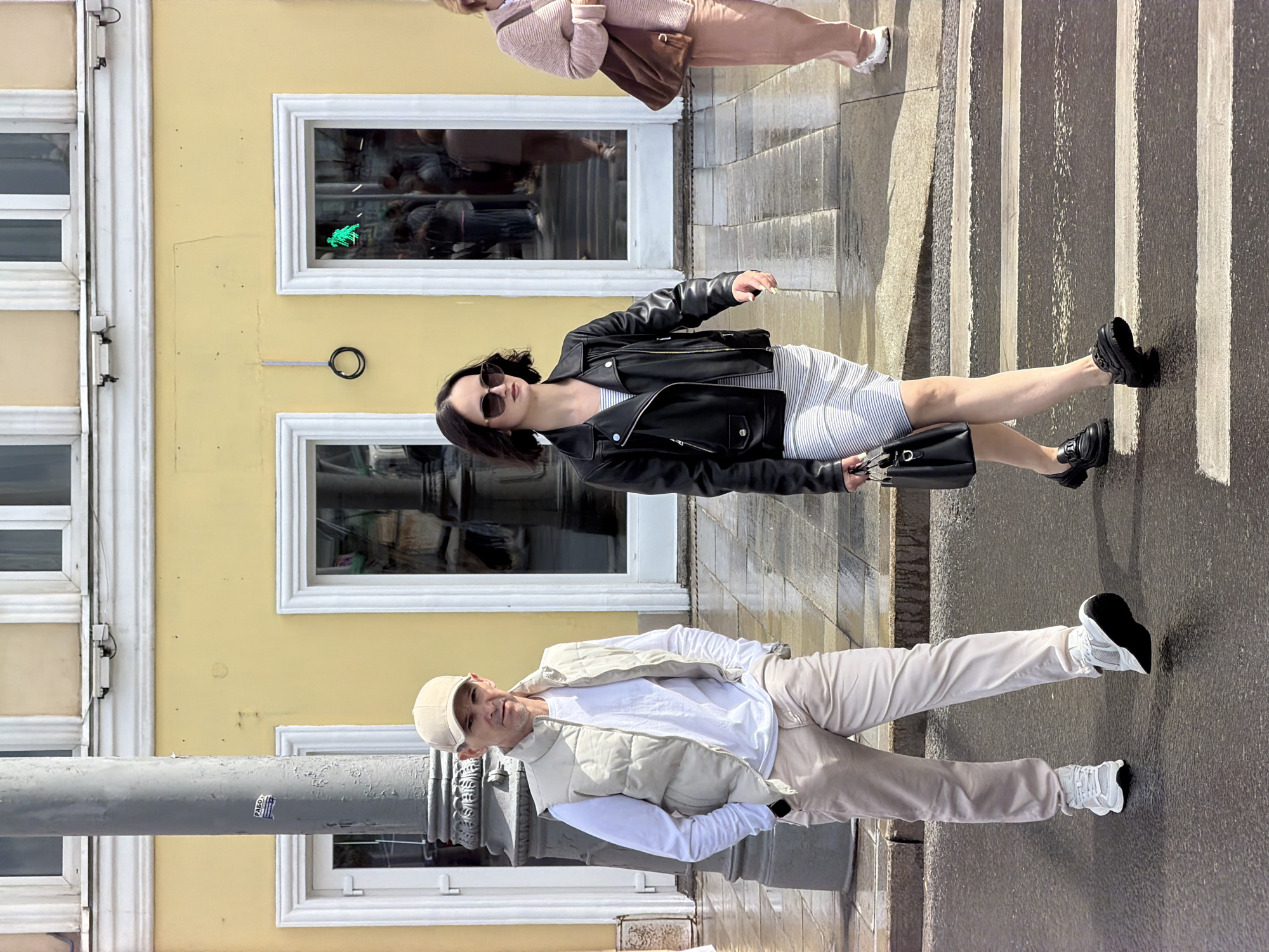 The image shows three people on a street in Moscow, Russia. The background is a light yellow building with white window frames. The image is taken at an angle, making it appear as if the people are walking sideways.
The woman in the middle is wearing a striped dress, a black leather jacket, sunglasses, and black loafers. She is carrying a black purse. The man at the bottom is wearing a beige cap, a white long-sleeve shirt, a beige vest, and beige pants. He is also wearing white sneakers. The person at the top is wearing a pink sweater and pink pants, and carrying a brown bag.
They are walking on a street with a zebra crossing. A gray pole is in the lower-left corner, and the street appears to be wet. There is a sticker on the pole.