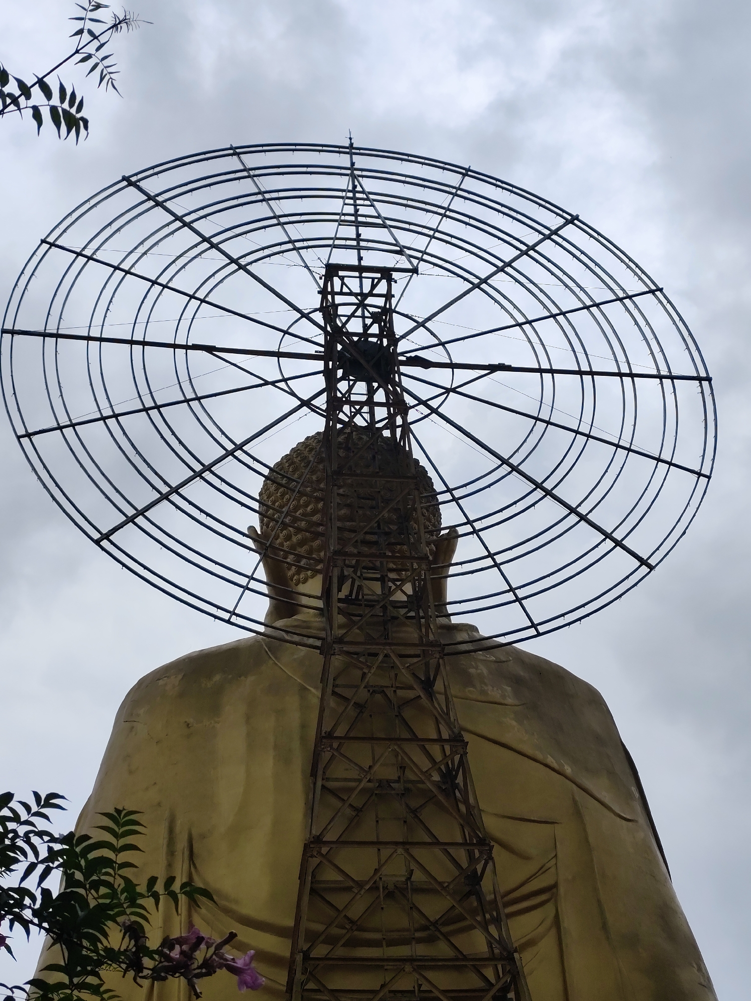 A low-angle shot of a golden Buddha statue under construction in Lâm Viên - Đà Lạt, Vietnam, viewed from behind. A large, circular metal frame structure with a central tower is being erected around the statue's head. The statue's back and head are visible, with the metal structure obscuring some details. Some foliage is visible in the lower left of the frame, and a small leafy branch is at the upper left. The sky is overcast.