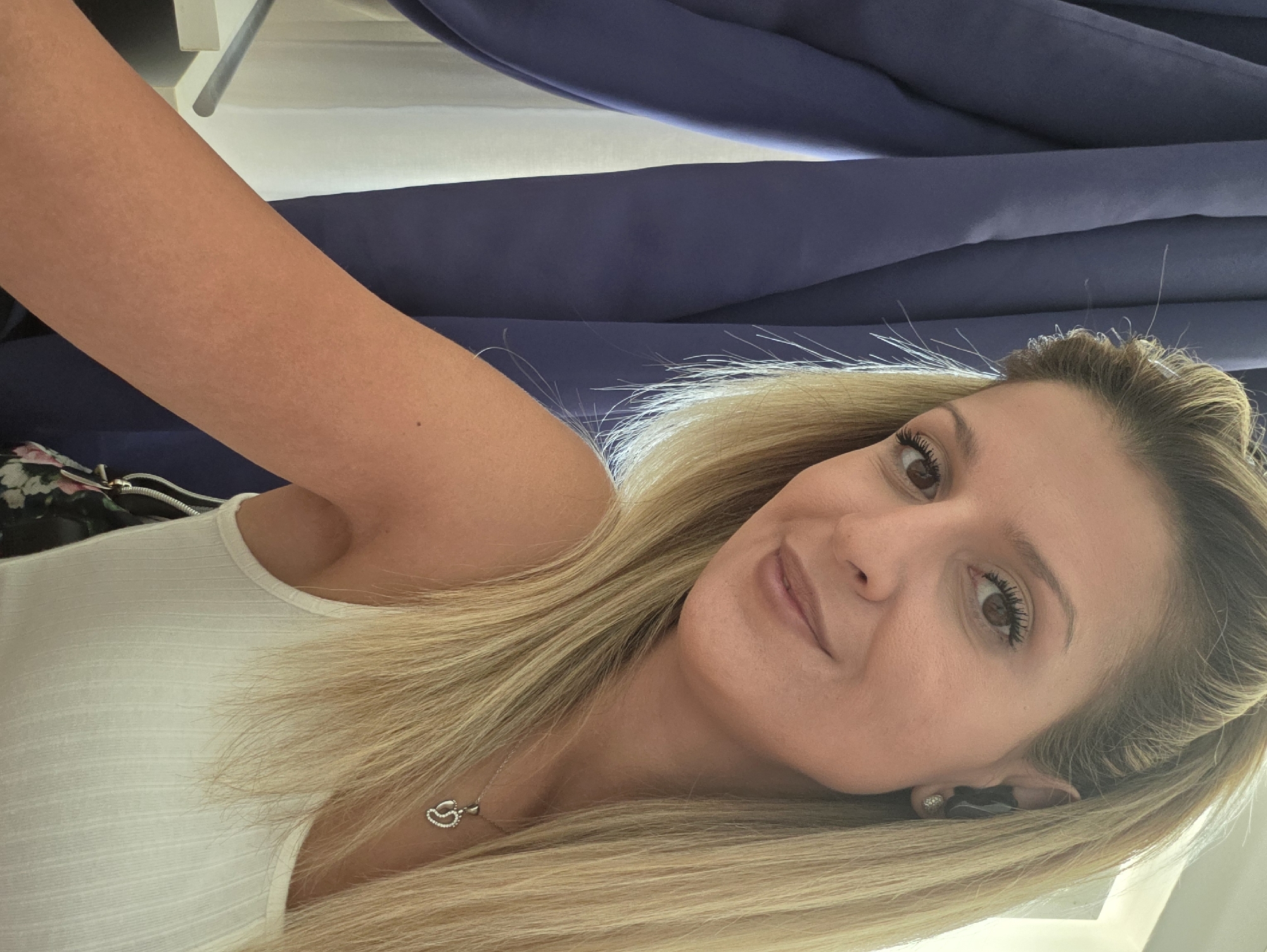 A woman with blonde hair and brown eyes is pictured from the chest up, taking a selfie indoors. She is wearing a white tank top and a silver necklace with a heart-shaped pendant. She is smiling and looking at the camera. In the background, there are blue curtains and a white curtain. The setting does not provide any specific information to determine the location in Munich, Germany.