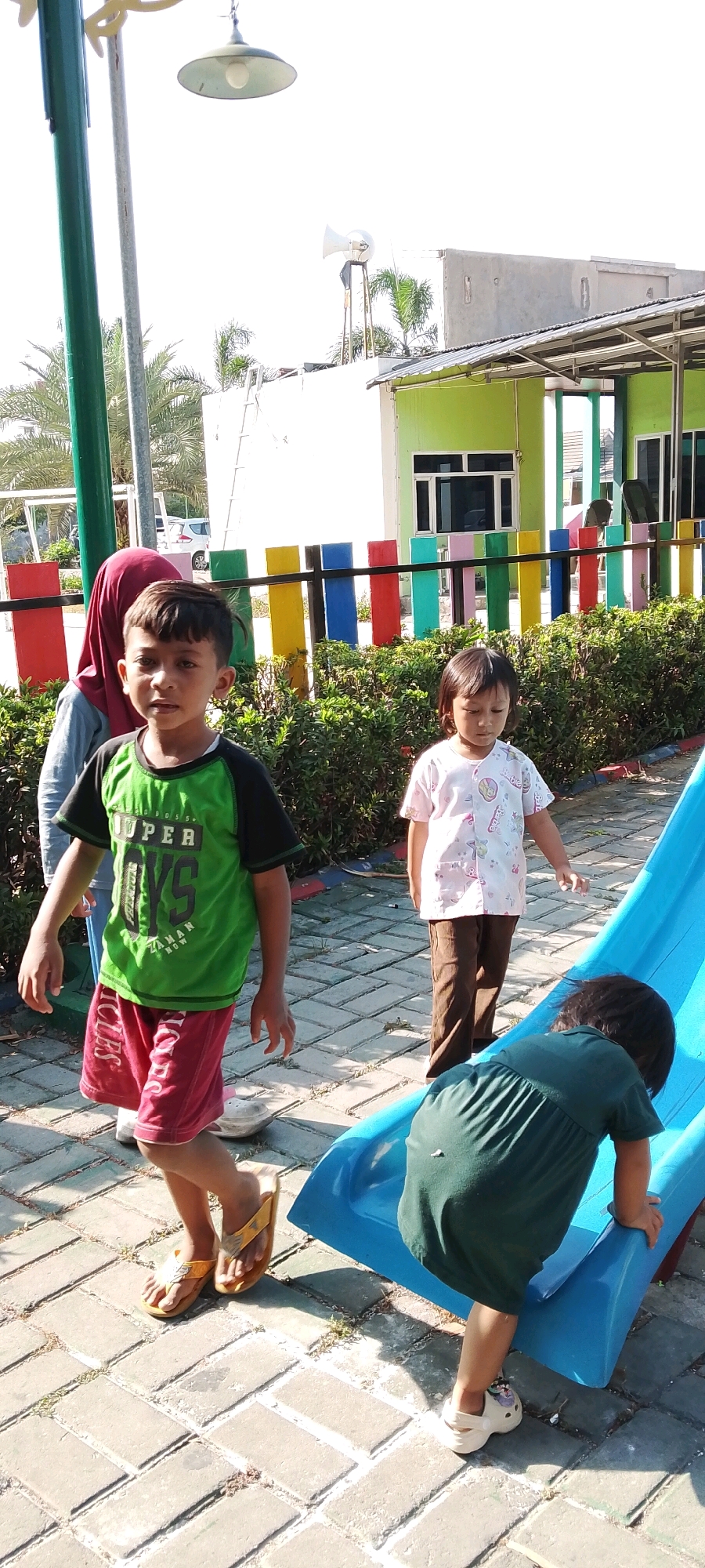 The image depicts an outdoor scene in Satriamekar, Indonesia, featuring four children and a playground area. In the foreground, a young boy in a green t-shirt with black sleeves and red shorts is standing and looking towards the camera. To his right, a younger girl in a light-colored, patterned shirt and brown pants is looking down. Further to the right, another young child wearing a dark green dress is on hands and knees, positioned at the bottom of a bright blue slide. A fourth child, partially visible in the background, is wearing a red headscarf. The children are on a paved walkway with a colorful fence and dense green bushes behind them. A building with a green facade and a corrugated metal roof is visible in the background, along with palm trees and a bright, possibly overcast sky. A street lamp with a wide shade is visible on the left side of the frame, and a white speaker horn is mounted on a pole above the building. The overall scene suggests a public park or playground.