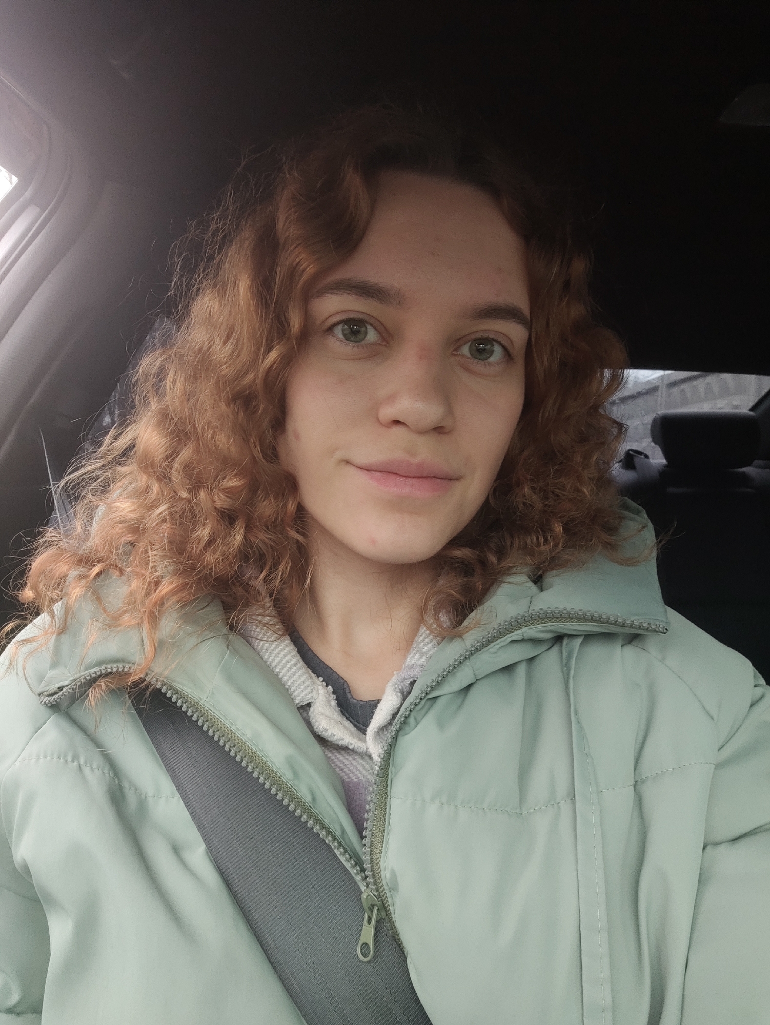 A woman with curly red hair is sitting in a car, wearing a light green puffer jacket with the seatbelt fastened. The car's interior is mostly dark, with the black ceiling visible. The woman has fair skin and green eyes. Outside the window, a blurry urban scene is visible, suggesting the location is within a city, presumed to be in Unknown, Unknown. The lighting indicates it might be daytime but overcast.