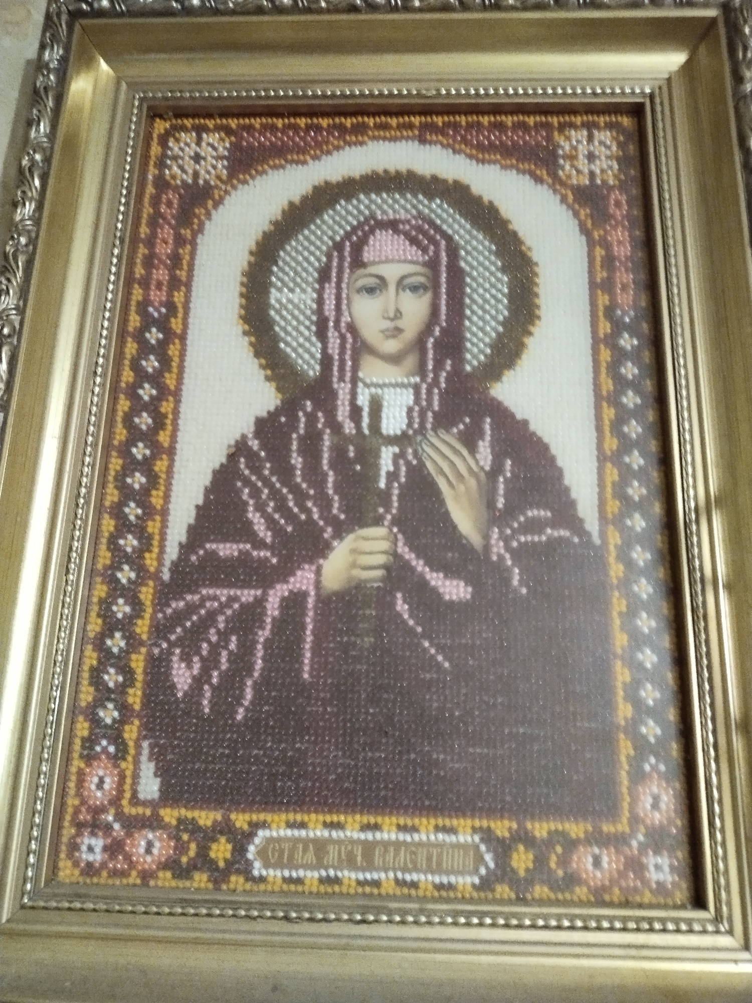 This media file presents a framed religious icon, likely a work of beadwork or intricate embroidery, found in Horishni Plavni, Ukraine. The central subject is a serene female saint, depicted frontally with a pale complexion and a gentle, solemn expression. She wears a pink head covering (maphorion) and dark, flowing robes, adorned with lighter pink patterns that suggest folds in the fabric. A luminous halo, intricately formed with white and silver beads, encircles her head. Her left hand gently holds a thin, staff-like object, possibly a cross or lily, while her right hand rests across her chest in a gesture of piety.
The icon features a richly patterned border in hues of brown, gold, and white, complementing the main figure. The entire artwork is enclosed within an ornate, gleaming golden frame with detailed carvings, adding to its devotional aesthetic.
At the bottom of the icon, Cyrillic text is clearly visible, reading "СТ.А ВЧ. ВАЛЕНТИНА". This translates to "Saint Great Martyr Valentina," identifying the saint depicted. No specific activity is shown; the image is a static, devotional representation. The beadwork technique creates a tactile and shimmering texture across the entire piece, catching the light and giving the icon a vibrant, almost three-dimensional quality.