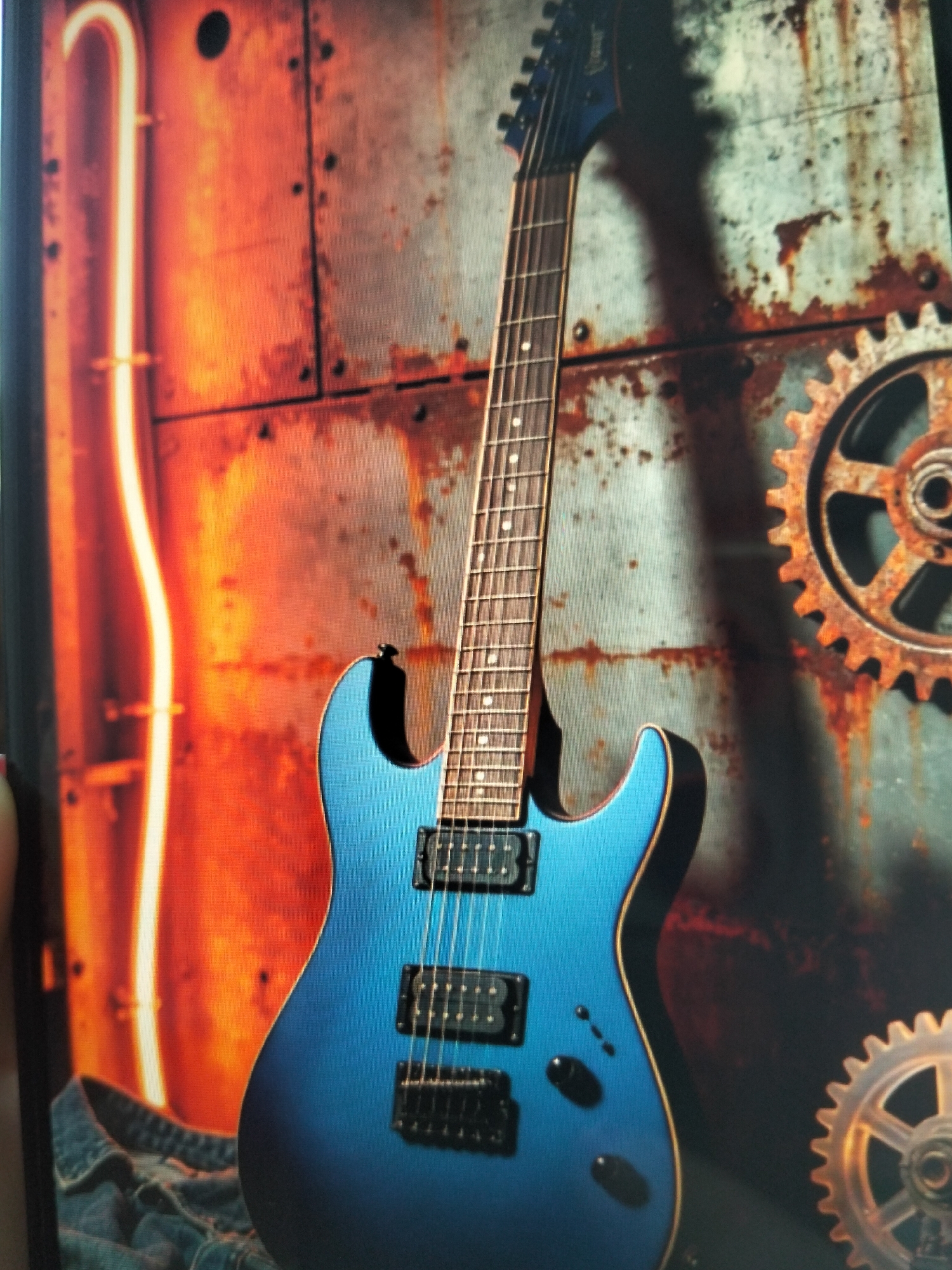 A blue electric guitar stands vertically in the foreground against a backdrop of a rusted metal wall with industrial gears. To the left, a vertical orange-lit structure is visible. The guitar features black pickups, a maple fretboard with dot inlays, and a dark headstock. Denim fabric is draped around the base of the guitar. The scene appears to be a studio or a staged setting, with no discernible indication of its location in Payagaboh, Indonesia.
