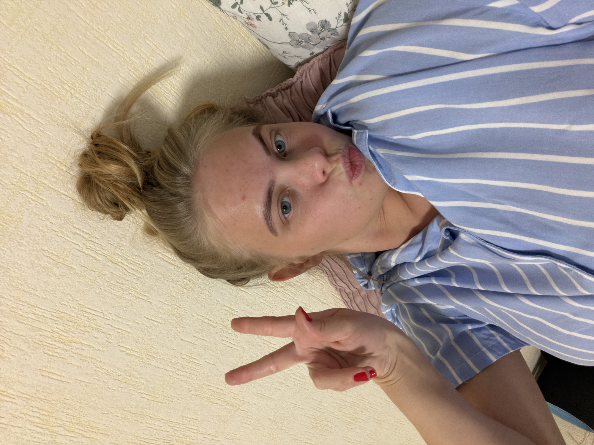 A woman with fair skin and blonde hair tied up in a bun is lying on a light-colored surface. She is wearing a blue and white striped shirt and making a kissing face. Her left hand is raised, showing a peace sign. Her fingernails are painted red. Part of a pillow with a floral pattern is visible behind her head. The background is a textured light-colored surface.
The location in the city of Izhevsk, Russia is unable to be determined.