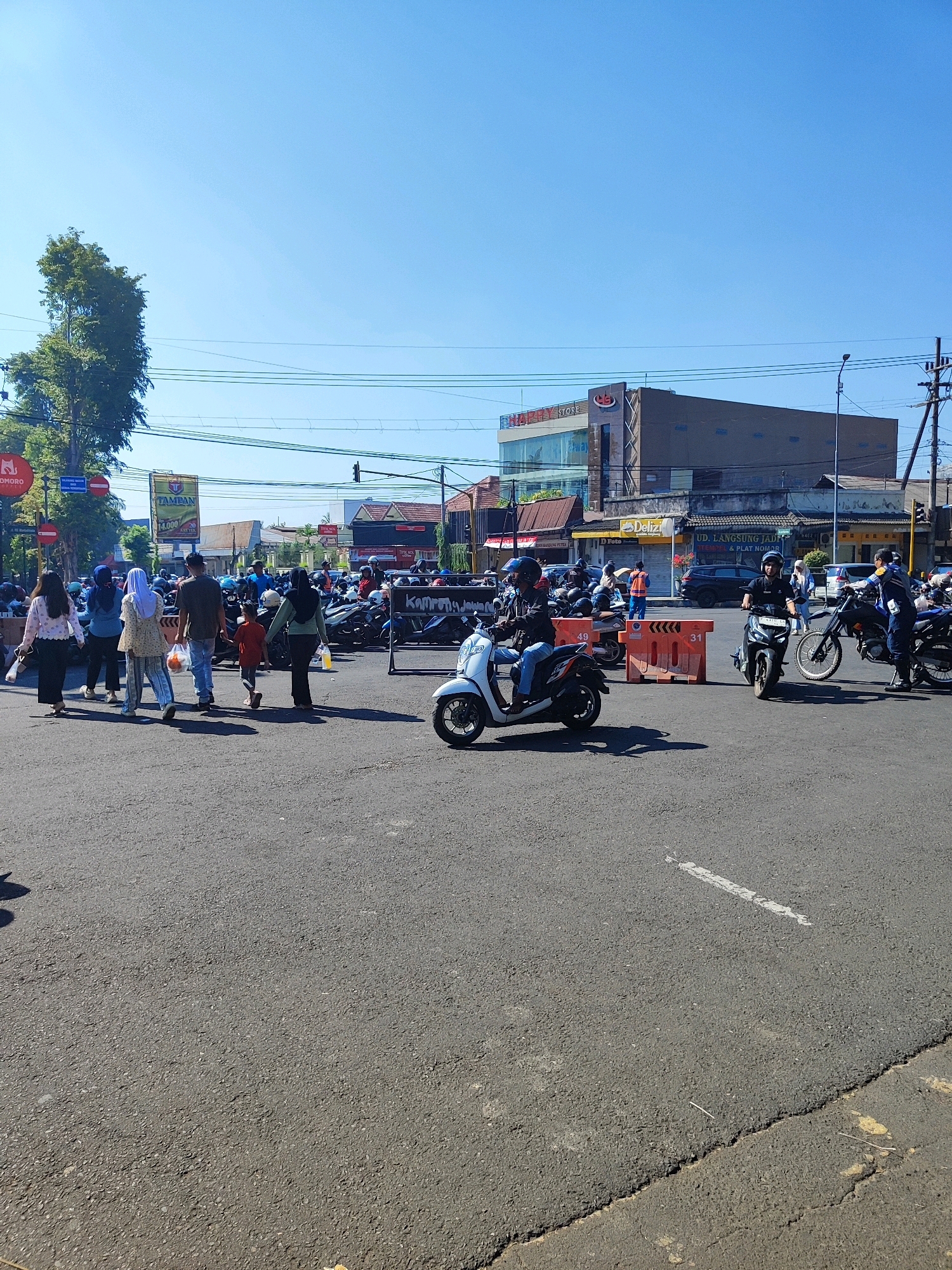 The image captures a street scene in Bondowoso, Indonesia, on a bright, sunny day with a clear blue sky. The foreground shows an asphalt road with white lane markings. In the mid-ground, several people are walking across the street, some carrying shopping bags, while others are on motorcycles. A young child is walking with an adult. Orange traffic barriers are placed on the road. In the background, there are several buildings, including what appears to be a store with a sign that says "HAPPY STORE". A large tree with dense foliage is visible on the left side of the image. A busy parking area filled with motorcycles is also present. Traffic lights and power lines are visible overhead. The scene appears to be a typical urban environment with moderate pedestrian and vehicle traffic.
