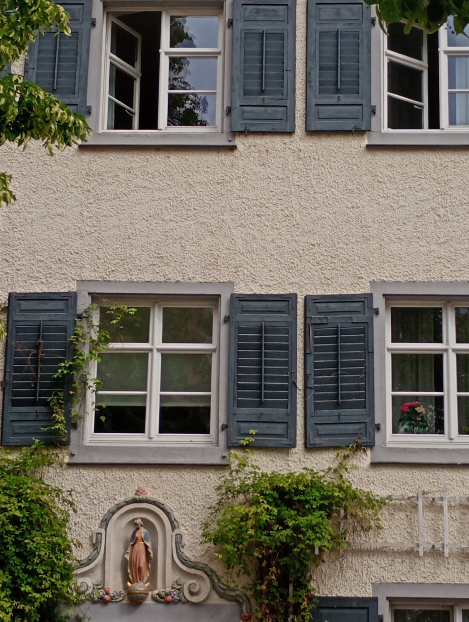 The image shows the exterior of a building with a stucco facade, likely in Meersburg, Germany. Multiple windows with dark blue shutters are visible. Some shutters are partially open, revealing the interiors. The upper windows have white frames and are divided into multiple panes. The left-most upper window is open. Green foliage from trees and vines partially obscures some of the windows and shutters. Below the windows, a decorative niche contains a statue of a woman in flowing robes. The niche is framed by an arched decorative element and flanked by floral motifs. A metal trellis structure is visible on the right side of the facade. The overall impression is of an older building with architectural detailing and a natural, overgrown aesthetic.