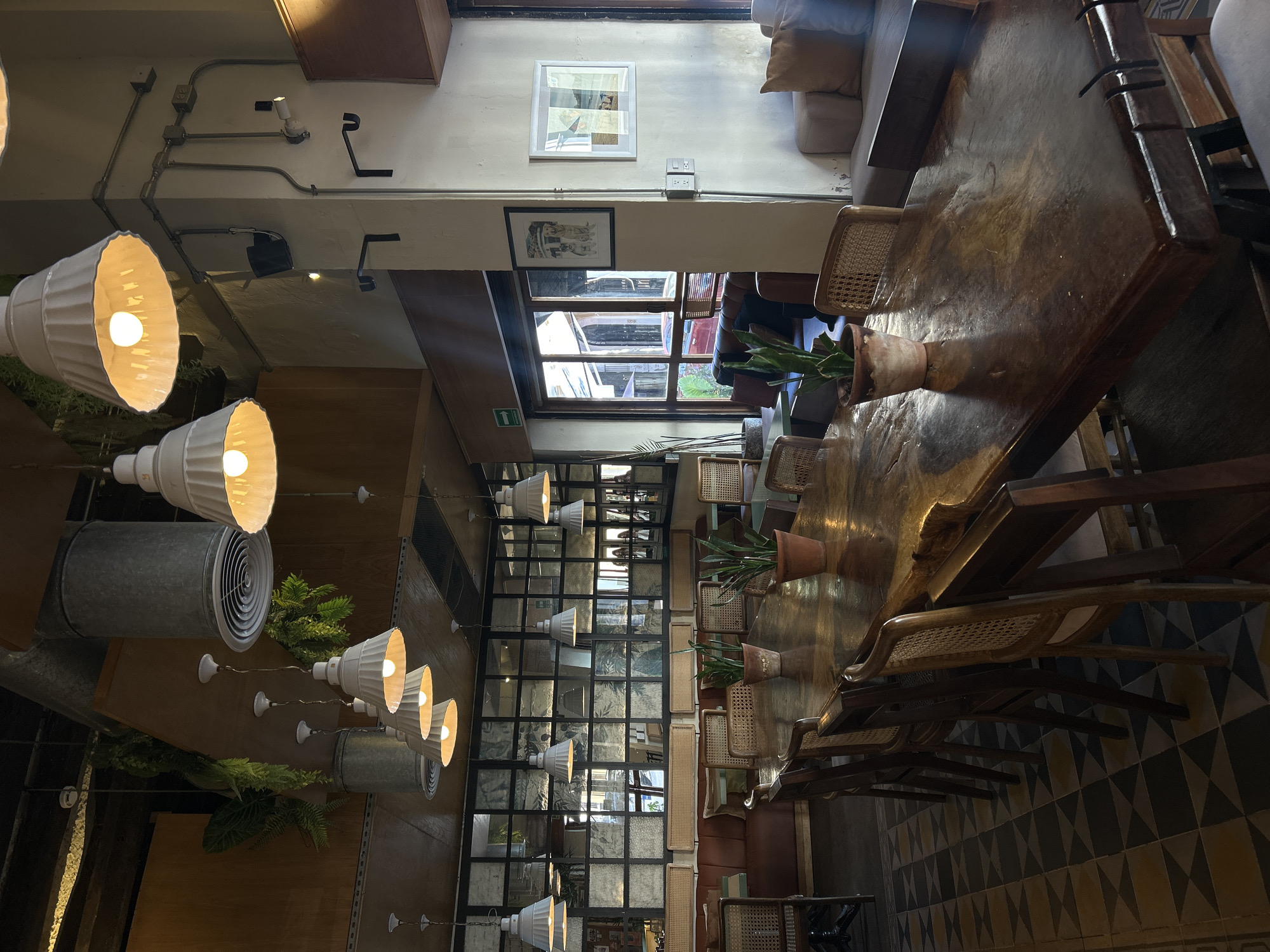 The image presents the interior of a stylish restaurant or cafe in Mazatlán, Mexico, characterized by a blend of industrial and rustic-chic design elements. The central feature is a long, dark, polished wooden communal table, capable of seating numerous guests. Along its length are arranged matching wooden chairs with woven cane backs and seats, some appearing to have cushions. Several potted green plants in terra cotta pots are placed at intervals on the table, contributing a natural touch to the decor.
The ceiling exposes industrial-style electrical conduits and features multiple large, white, bell-shaped pendant lights, which cast a warm, inviting glow. Smaller spotlights and prominent circular metallic ventilation ducts are also visible. Cushioned benches or sofas line one wall, offering additional seating options.
A large multi-paned window provides natural light and a view of the outside, where vehicles and greenery can be partially seen. On an opposite wall, a grid-patterned glass partition creates reflections of the interior space. Framed pictures hang on the walls, and a visible green "EXIT" sign with an arrow pointing left indicates an emergency exit. The floor is adorned with distinctive geometric tiles in patterns of dark blue, yellow, beige, and white.
The scene is devoid of people, suggesting it was captured during off-peak hours or when the establishment was quiet, lending a peaceful and serene ambiance to the space. Despite the natural light streaming in, the interior lights are on, enhancing the warm and inviting atmosphere. The overall impression is one of a thoughtfully designed space ready to host patrons for dining or socializing.