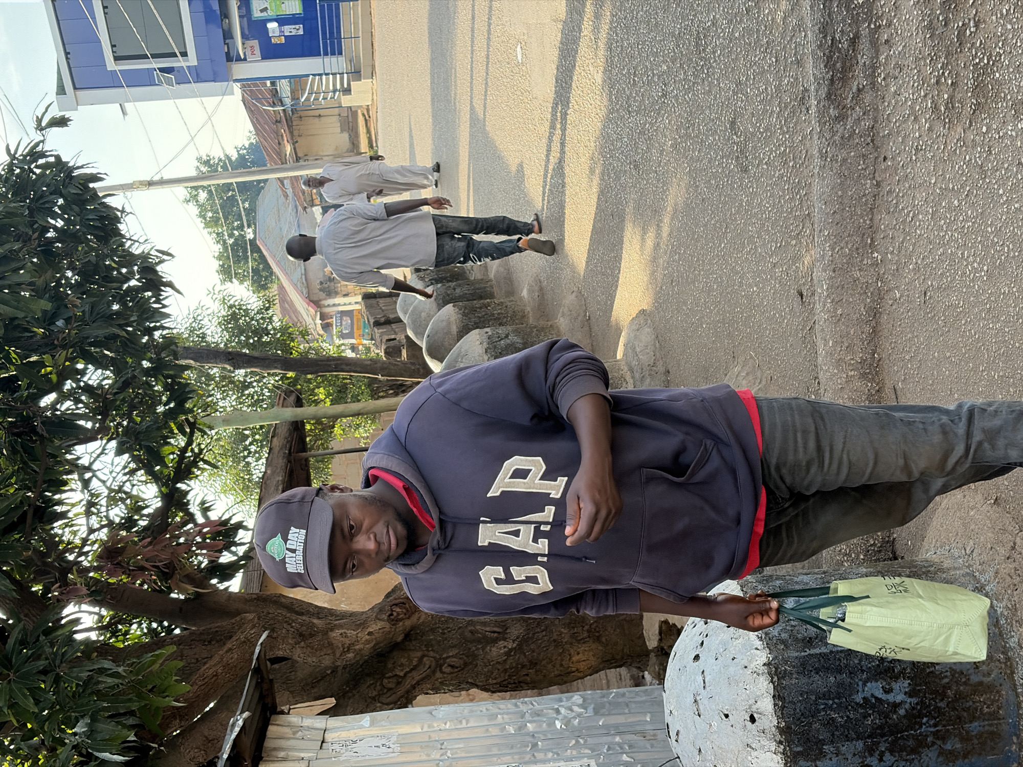 The image shows a man walking on a paved road in the city of Jos, Nigeria. The man is wearing a navy blue Gap hoodie with a red shirt underneath, dark jeans, and a black baseball cap with "MAY DAY CELEBRATION" printed on it. He is carrying a light green reusable shopping bag in his left hand. In the background, two other men are walking away from the camera, and a blue building with "POS" signage is visible. The scene appears to be set outdoors in a street environment, possibly during daytime given the lighting. There are trees and some structures indicating an urban setting.
