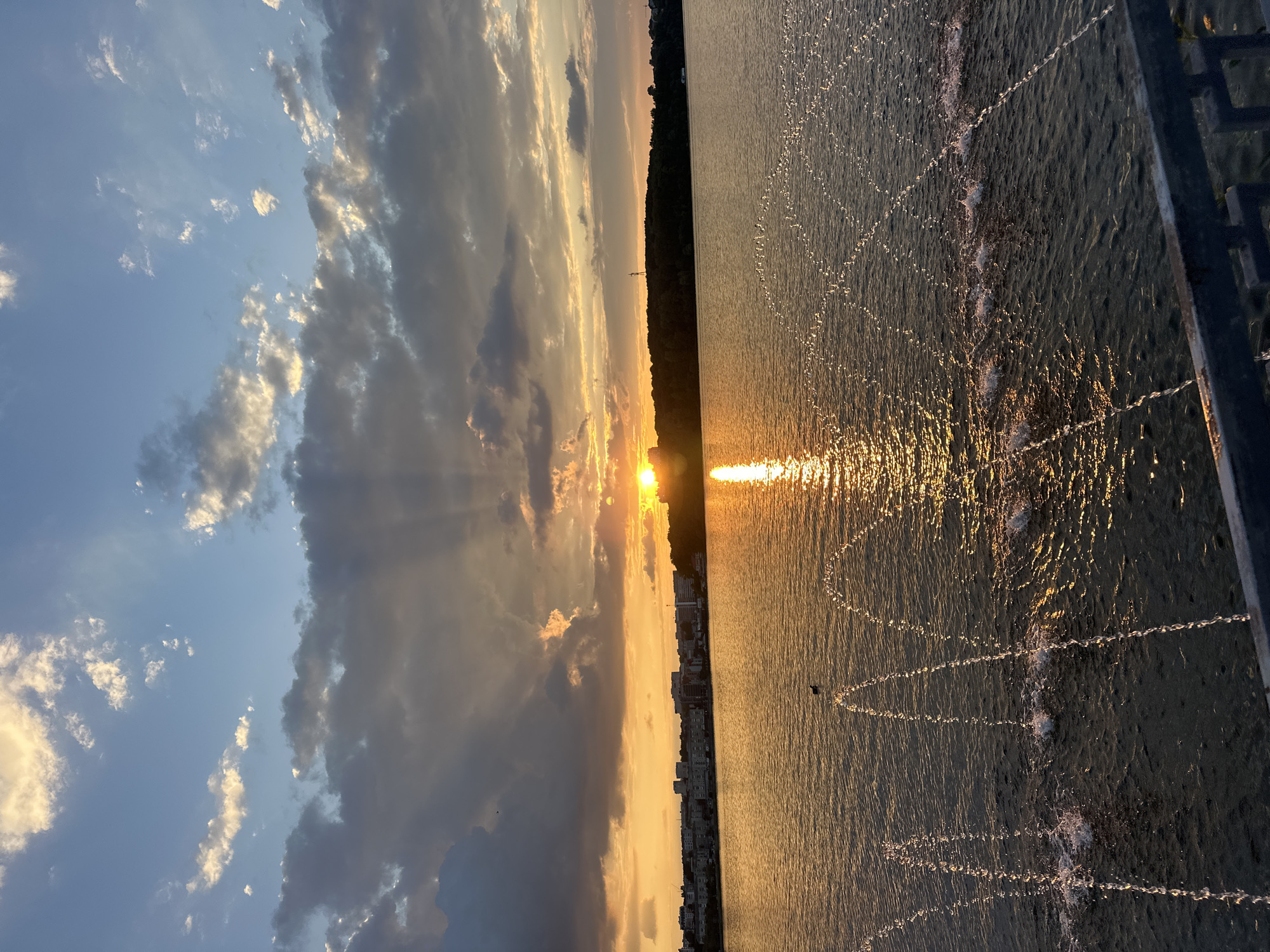 This is a wide shot of a sunset over the Ternopil pond in Ternopil, Ukraine. The sun is low on the horizon, casting a golden glow across the water. The sky is filled with dramatic clouds, some of which are illuminated by the setting sun. In the foreground, water jets from a fountain create arcs of spray. A small bird can be seen on the water, and a cityscape is visible in the distance, along with a treeline. The overall mood is serene and picturesque.