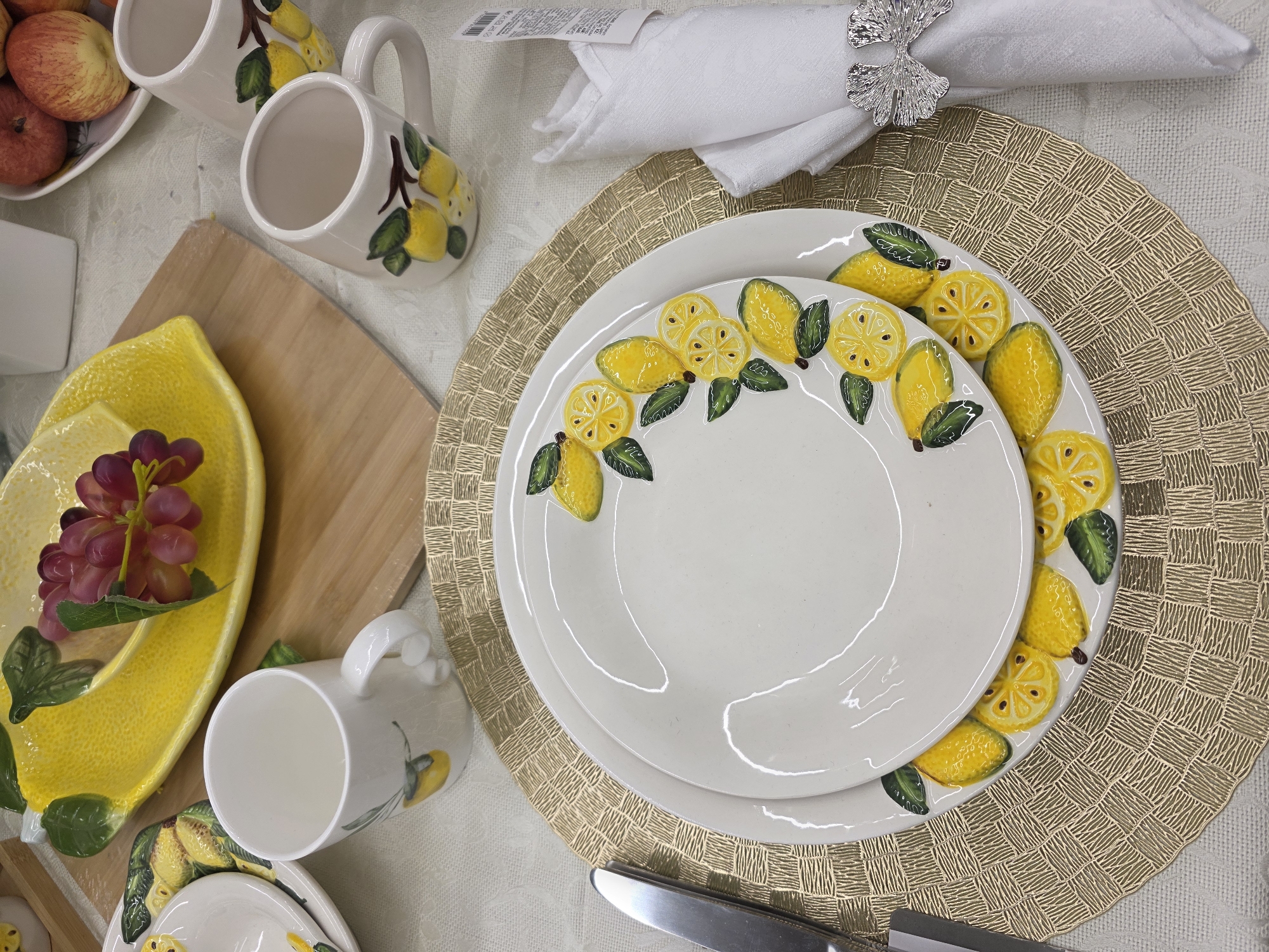 This image depicts a detailed table setting in an indoor environment, potentially a retail display or dining area, located in Chaikovsky, Russia. The scene centers on a table covered with a white, subtly patterned tablecloth.
A prominent place setting is visible in the foreground, featuring a round, gold-colored placemat with a woven texture. Stacked on this placemat are two white ceramic plates: a larger dinner plate beneath a smaller salad plate. Both plates are adorned with a raised, hand-painted design of yellow lemons and green leaves arranged in a semi-circular pattern along their rims. To the left of the plates, a silver knife is positioned. To the right, a white linen napkin is neatly folded and held by a silver bow-shaped napkin ring. A price tag or label is attached to the napkin.
Further back, two white ceramic mugs, each decorated with yellow lemons and green leaves, are present. One mug is positioned to the left of the main place setting, and another is visible in the upper right quadrant. A large, bright yellow, lemon-shaped ceramic serving dish, containing a sprig of artificial purple grapes, rests on a light brown wooden cutting board positioned towards the upper left. In the upper right background, a white bowl filled with red apples is partially visible. No individuals or actions are depicted in the scene.