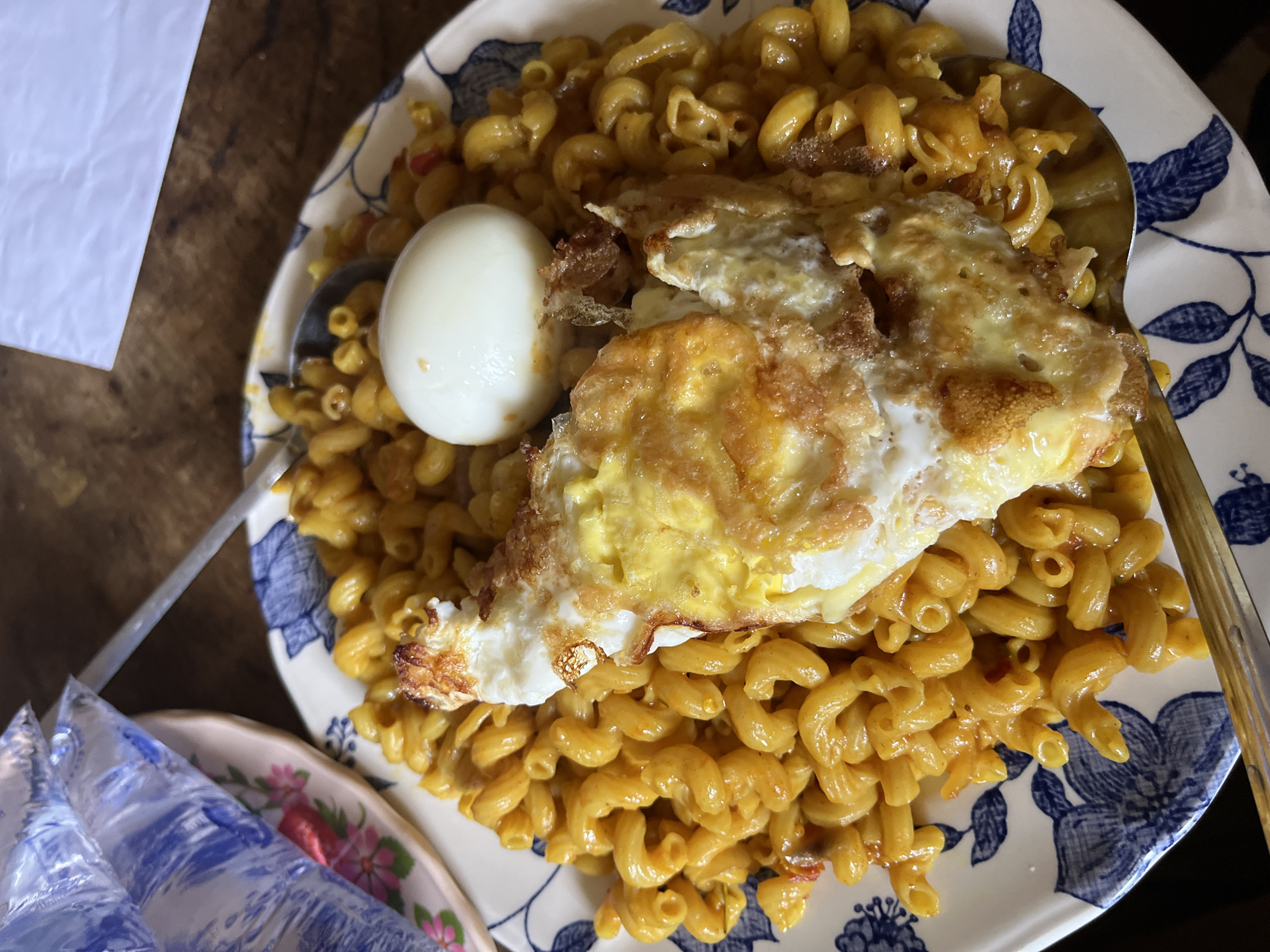 A plate of food, likely macaroni with sauce and a fried egg, is presented on a wooden surface. The plate has a blue and white floral pattern. To the left of the plate, a pink and white floral saucer holds two plastic-wrapped items, possibly water pouches. A fork rests on the plate, partially submerged in the macaroni. The scene appears to be an indoor setting, possibly a home or a casual dining establishment. The lighting suggests it is daytime. There are no people visible in the image.