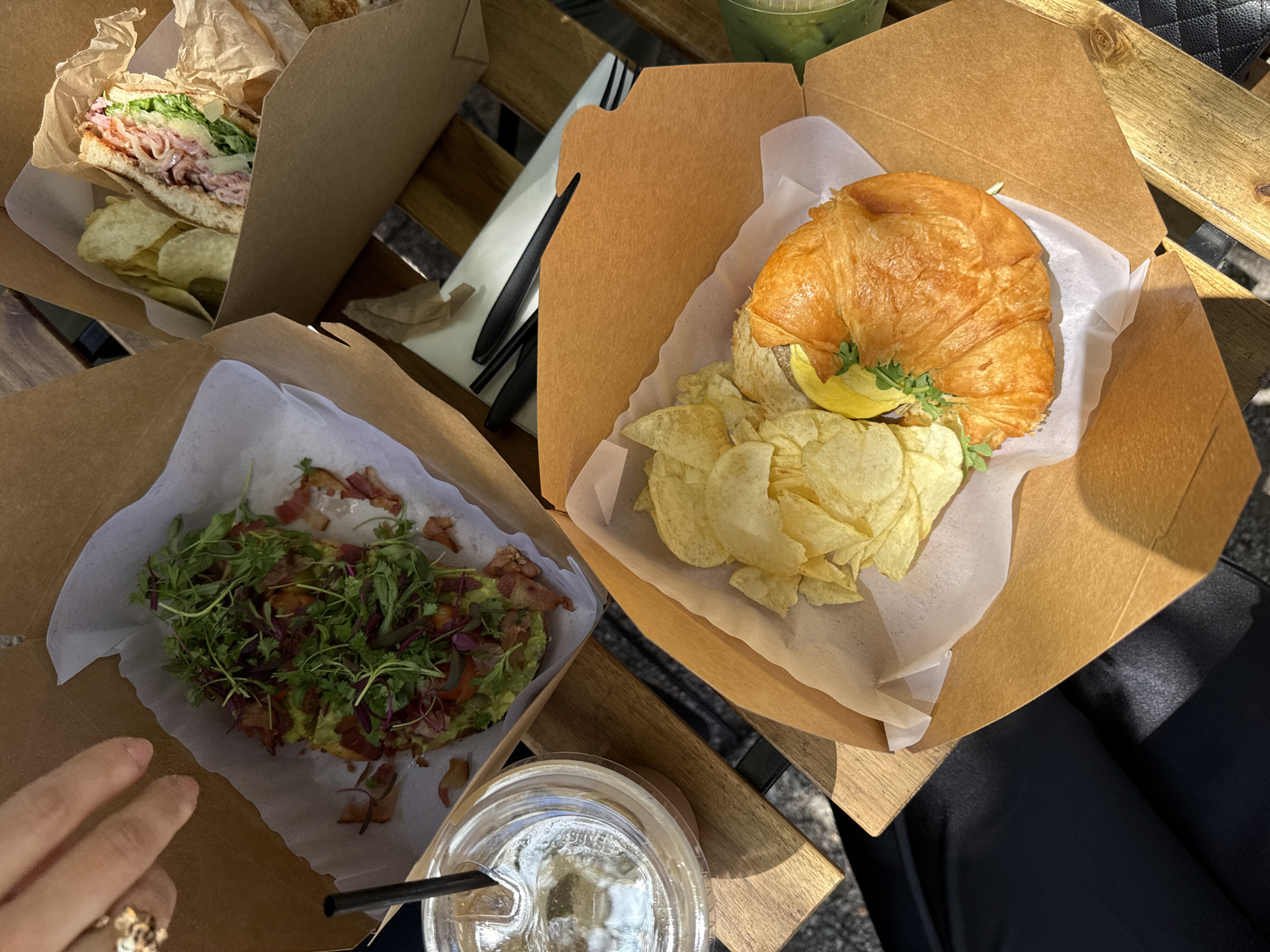The image shows a table with three food containers and two drinks. The containers hold a sandwich with chips, avocado toast, and a croissant with chips. The drinks are in clear plastic cups, one with a black straw. A hand with a ring is visible near the avocado toast. The setting appears to be outdoors on a wooden table. The image was taken in Chula Vista, California.