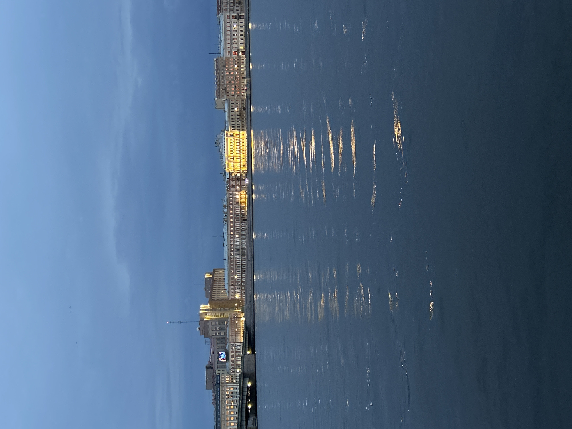 The image shows a wide body of water in the foreground, reflecting lights from a city skyline in the background. The sky is a deep blue, with some clouds visible, suggesting it is twilight or early evening. The city skyline features a series of buildings with illuminated windows, indicating that the city is active. A bridge is visible crossing the water, connecting different parts of the city. The water is choppy, with small waves creating ripples that catch the light. The overall impression is a serene urban landscape at dusk.