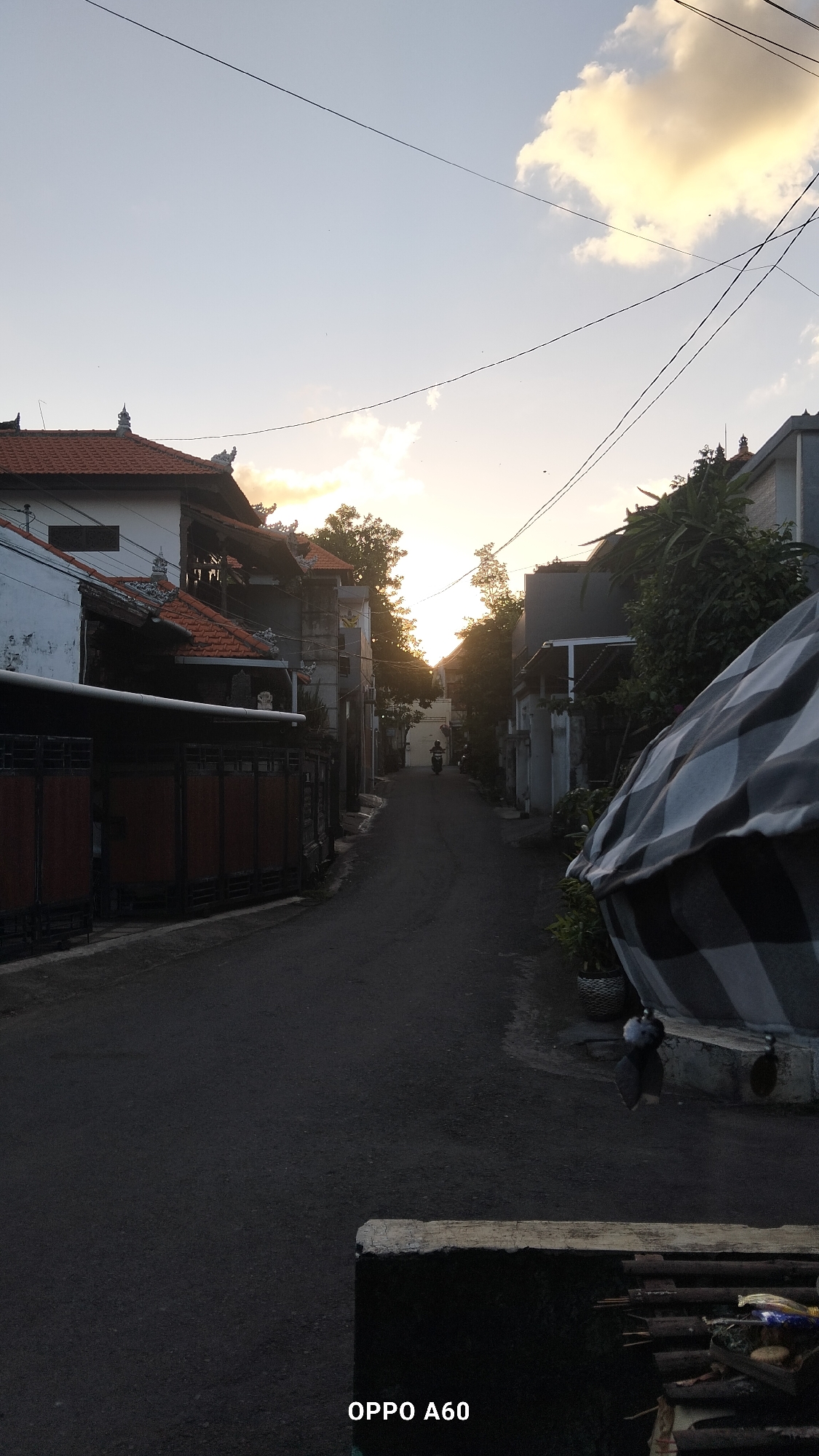 The image captures a street scene in North Kuta, Indonesia, during what appears to be sunset. The perspective is from a low vantage point, looking down a paved street lined with traditional Balinese-style houses. The architecture features distinctive tiled roofs with ornate details, and some walls are white or gray. On the left side of the street, a dark wooden gate is visible, suggesting a residential compound. On the right, there are more houses and lush green foliage.
The sky is a pale blue with scattered clouds tinged with yellow and orange from the setting sun, which is positioned towards the center of the frame, partially obscured by trees. A single individual on a motorcycle is traveling away from the camera down the street, the only discernible human presence. In the foreground, a portion of a black and white checkered tarpaulin is draped over an object, partially obscuring the lower right side of the image. There is also a pile of firewood and some other miscellaneous items in the immediate foreground, on the bottom edge of the frame. The overall atmosphere is serene, with a sense of quietude as day transitions into evening. A watermark at the bottom of the image reads "OPPO A60".