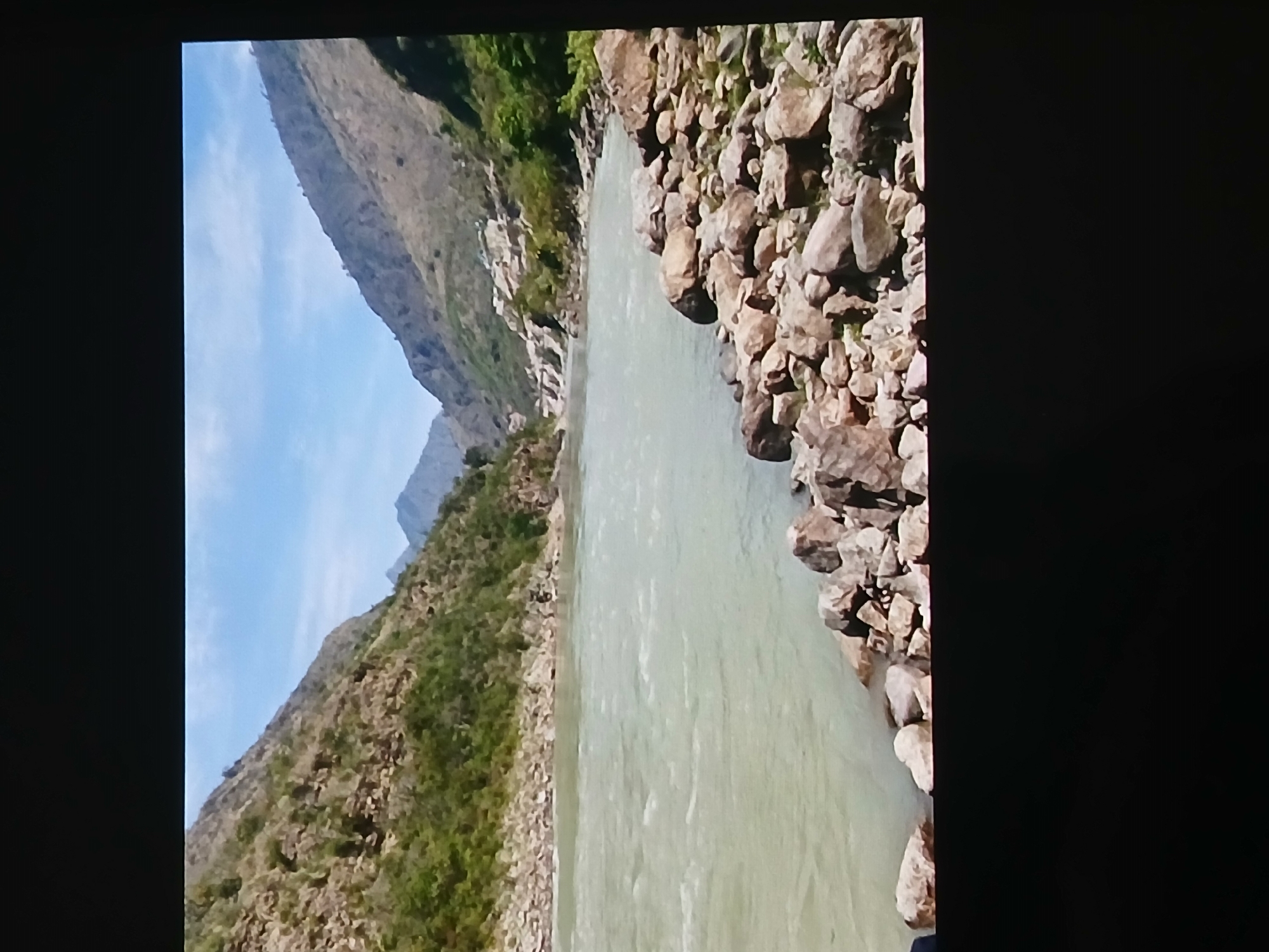 The image depicts a wide river flowing through a mountainous landscape. The river water appears murky and light in color. The river banks are lined with large rocks on one side and a vegetated slope on the other. Mountains rise on either side of the river, with a clear blue sky visible above. It is not possible to discern the location of the image as Bengaluru, India from the visual information provided.