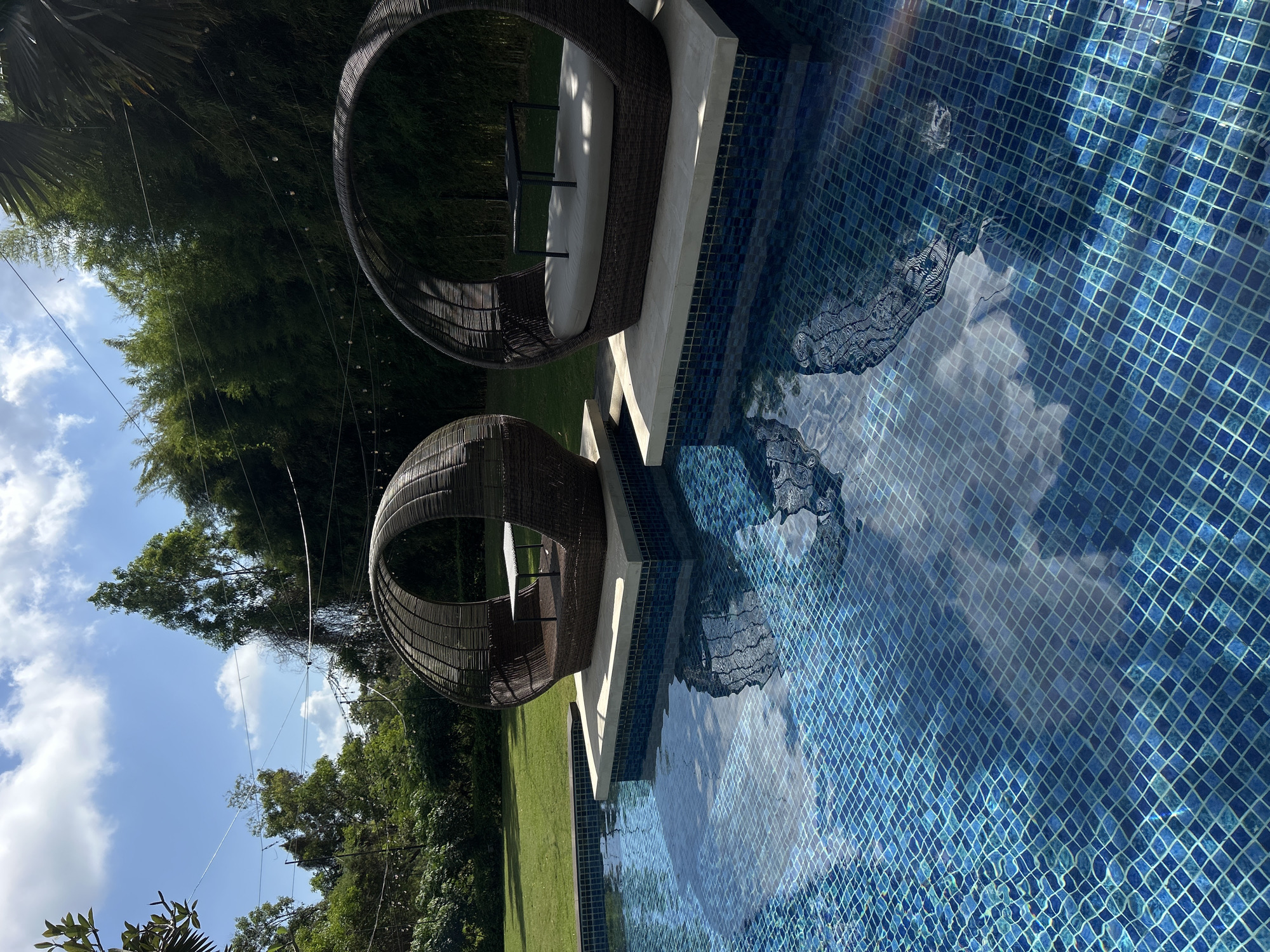 Two circular, woven loungers with integrated tables are situated on submerged platforms in a tiled swimming pool. The loungers are made of dark, woven material and feature light-colored cushions and dark tabletops. The pool is filled with clear blue water, reflecting the sky and surrounding environment. Behind the pool, a lush green lawn extends to a dense wall of bamboo trees. String lights are visible overhead, suspended between trees and poles. The sky is blue with scattered white clouds. The location is identified as Kuta Selatan, Indonesia. No people are present in the image.