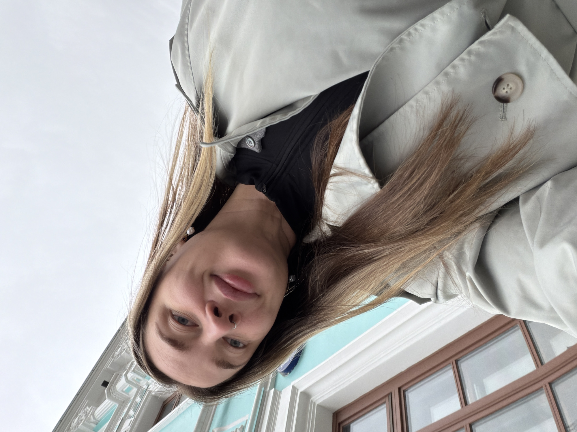 A low-angle shot of a woman, possibly a selfie, shows her face and upper body, framed by a light grey trench coat. She has fair skin, light brown hair with blonde highlights, and a nose ring. She is wearing a black garment underneath the coat. The background reveals a building with white architectural details and turquoise accents, along with brown-framed windows. The image was taken in the city of Moscow, Russia.