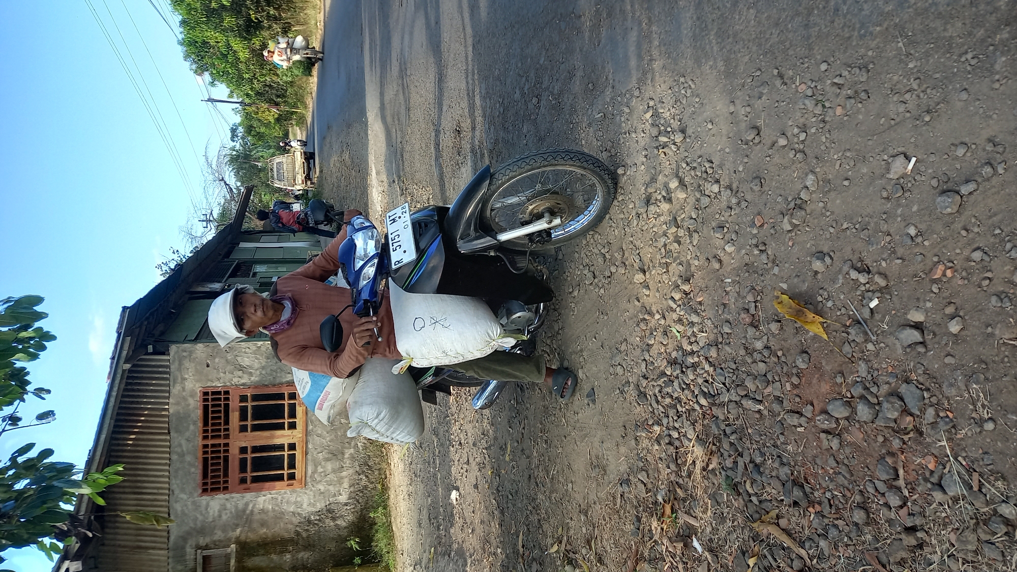 In Sidamulya, Indonesia, a man wearing a brown long-sleeved shirt, olive green pants, and a white head covering sits on a blue and black motorcycle. The motorcycle carries two large white sacks, one on each side. The man is on a dirt and gravel road in front of a weathered building with corrugated metal siding and a wooden-framed window. The background shows a paved road with other vehicles and lush greenery. The sky is clear and blue.