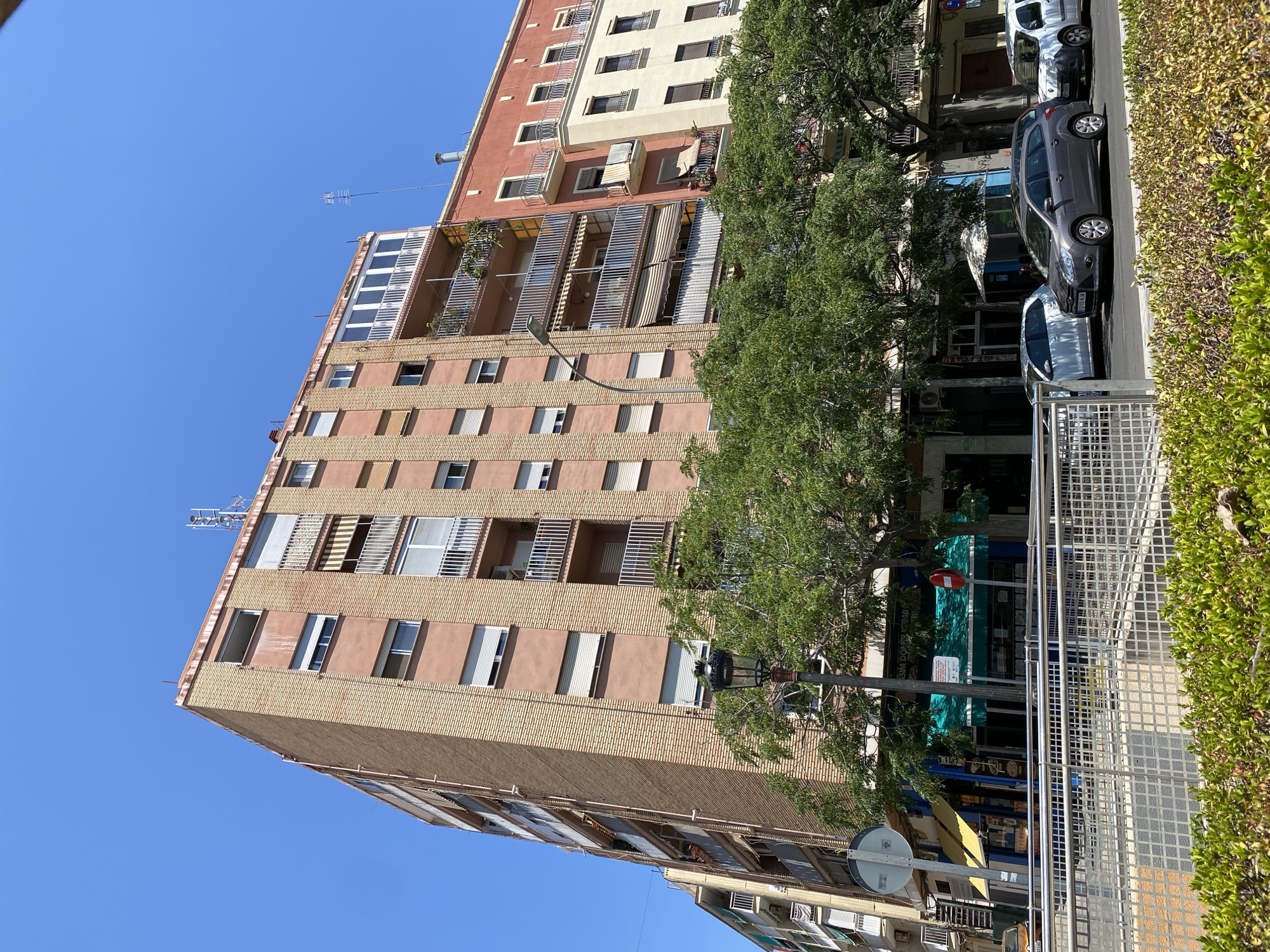 This is a photograph taken in València, Spain, on a clear, sunny day. The image features a tall, multi-story building with a light brown brick facade and reddish-pink upper floors. The building has numerous windows, many with white shutters, and several balconies with metal railings. Some balconies have plants, awnings, and chairs, suggesting residential use. An antenna is visible on the rooftop.
In front of the building, a vibrant green tree with dense foliage partially obscures the lower floors. A street lamp with a distinctive curved pole stands near the tree. Adjacent to the building, a sidewalk runs alongside parked cars. Three cars are visible: a dark gray hatchback, a white car, and a lighter-colored car further down the street. A metal fence runs along the edge of the sidewalk, bordering a green, leafy hedge.
The lower part of the photograph shows more of the building's facade and glimpses of storefronts with signs. One sign partially visible says "SIM DONER KEBAB". Another sign appears to be a red circular object on a pole. The overall scene depicts a typical urban streetscape.