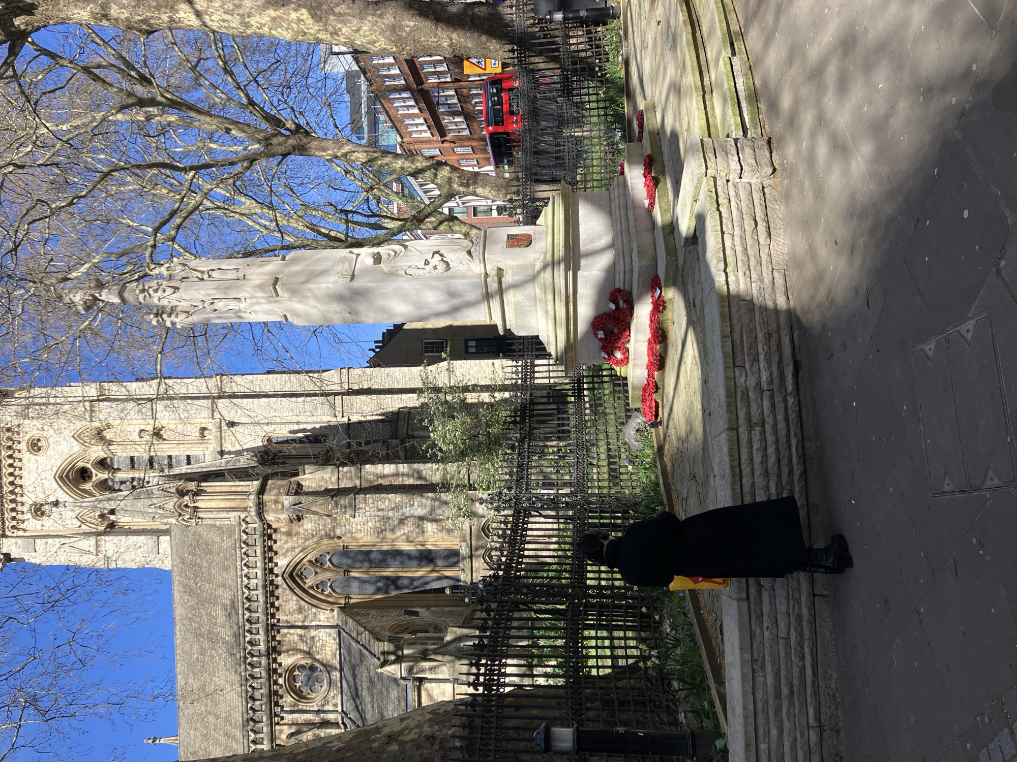 The image depicts a street scene in London, United Kingdom, specifically on Fleet Street, featuring St Dunstan-in-the-West church and the City of London School War Memorial. In the foreground, a single individual stands on a paved sidewalk with their back to the camera, wearing a dark, long coat, dark trousers, and black platform boots, holding a yellow bag in their left hand. They face a light-colored stone war memorial, which stands on a raised stone base behind a black wrought-iron fence. The memorial is adorned with multiple red poppy wreaths arranged around its base. Behind the fence and memorial is the Gothic-style stone church of St Dunstan-in-the-West, characterized by its ornate tower, arched windows, and detailed stonework. Bare-branched trees frame the scene on both sides and against a clear blue sky. In the background, beyond the church and trees, residential and commercial buildings are visible, including a red double-decker bus on a street. Shadows are cast across the paved ground, indicating a sunny day.